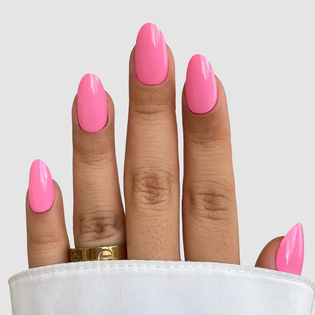 A hand with medium skin tone shows long, oval nails painted glossy pink using Sassy Saints - Brings The Salon Home's Sweet Beat Gel Wrap. The person wears a gold ring, and the nails emerge from a white sleeve against a light gray background.