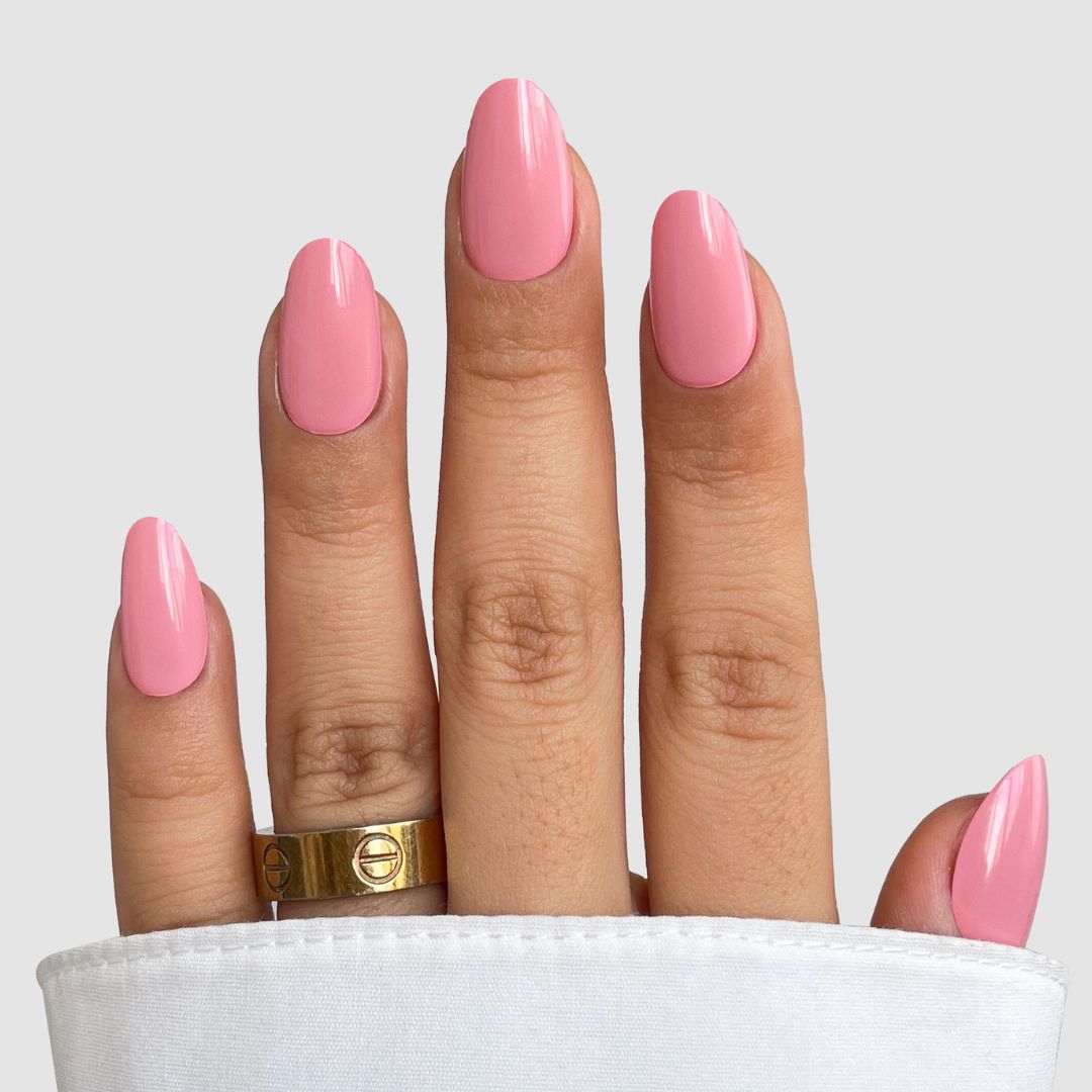 A hand with long, oval nails featuring Sassy Saints - Brings The Salon Home's Rose Tone Gel Wrap, painted glossy pink and wearing a thick gold ring. A white sleeve partially covers the hand against a light gray background.