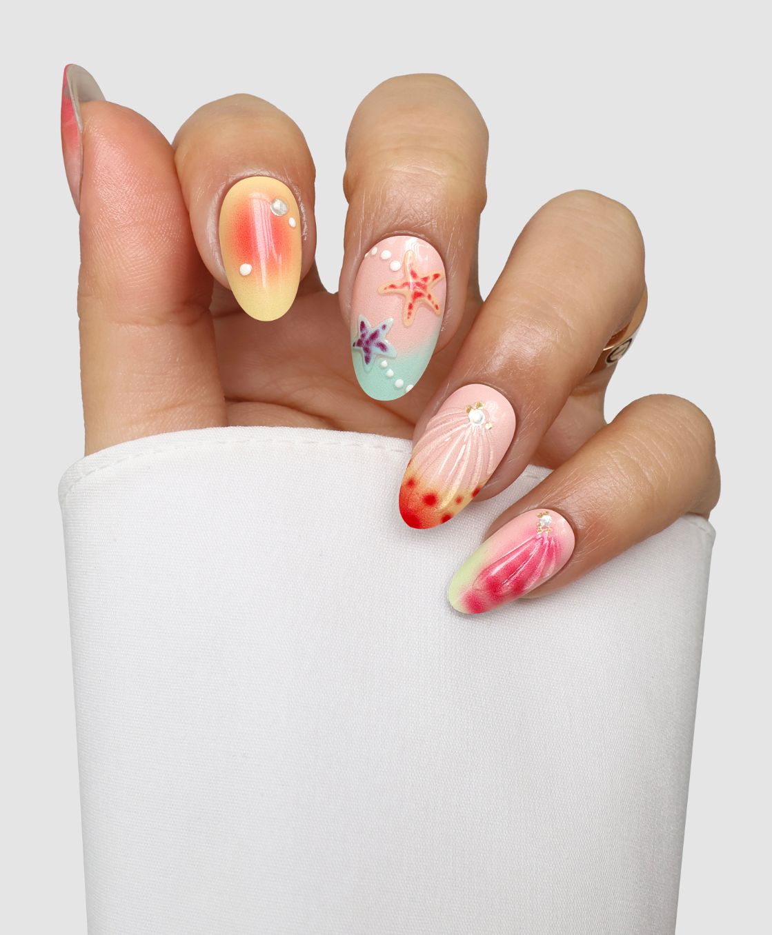 A hand with long, almond-shaped nails showcases Sassy Saints' Mermaid Mood (3D) Gel Wraps, featuring beachy 3D seashells, starfish, pearls, and pastel gradients. The nails pop against a backdrop of white fabric. Salon-quality art at home!.
