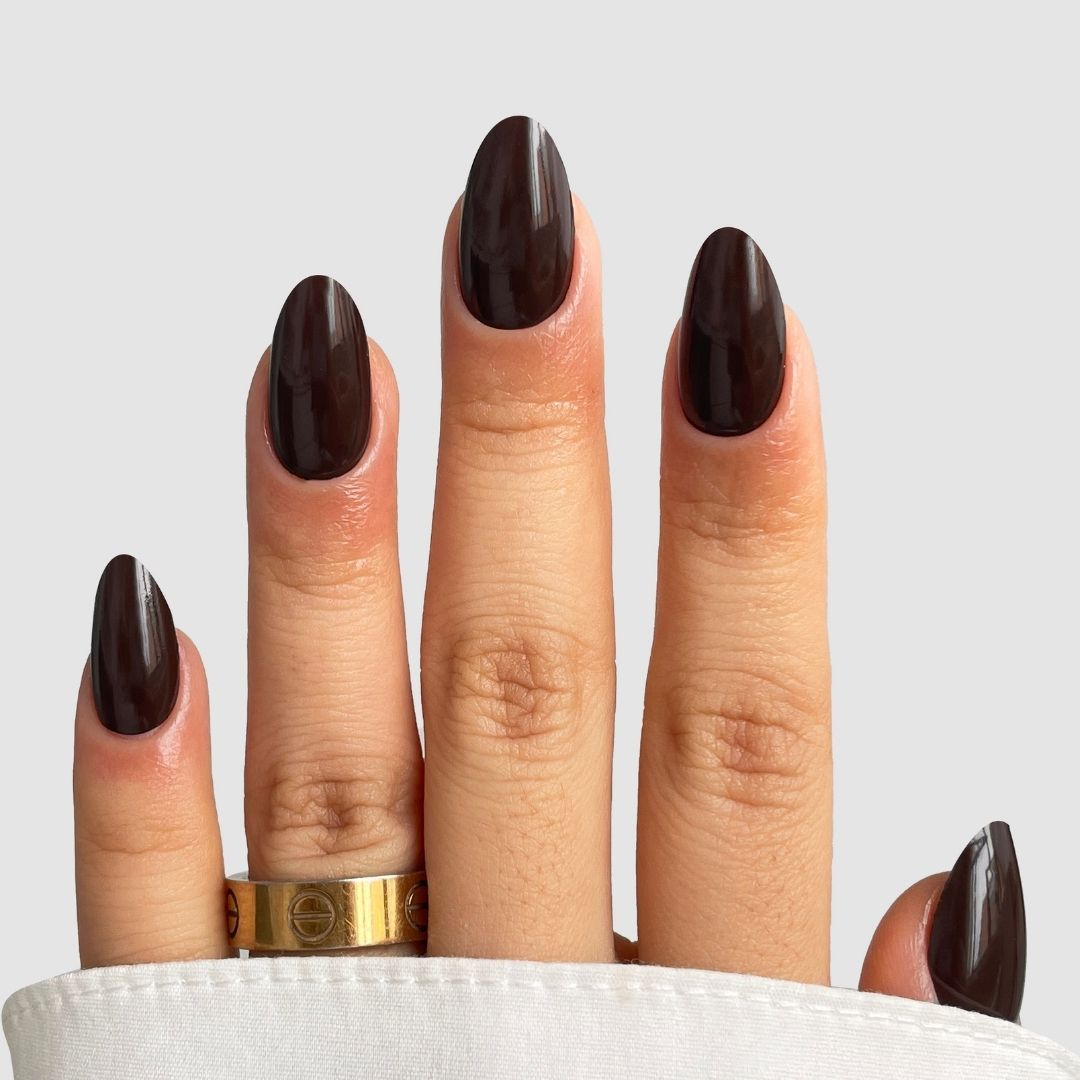 A hand with almond-shaped nails painted in Sassy Saints’ Cocoa Luxe (Dark Brown) polish, wearing a gold engraved ring, against a light gray background.