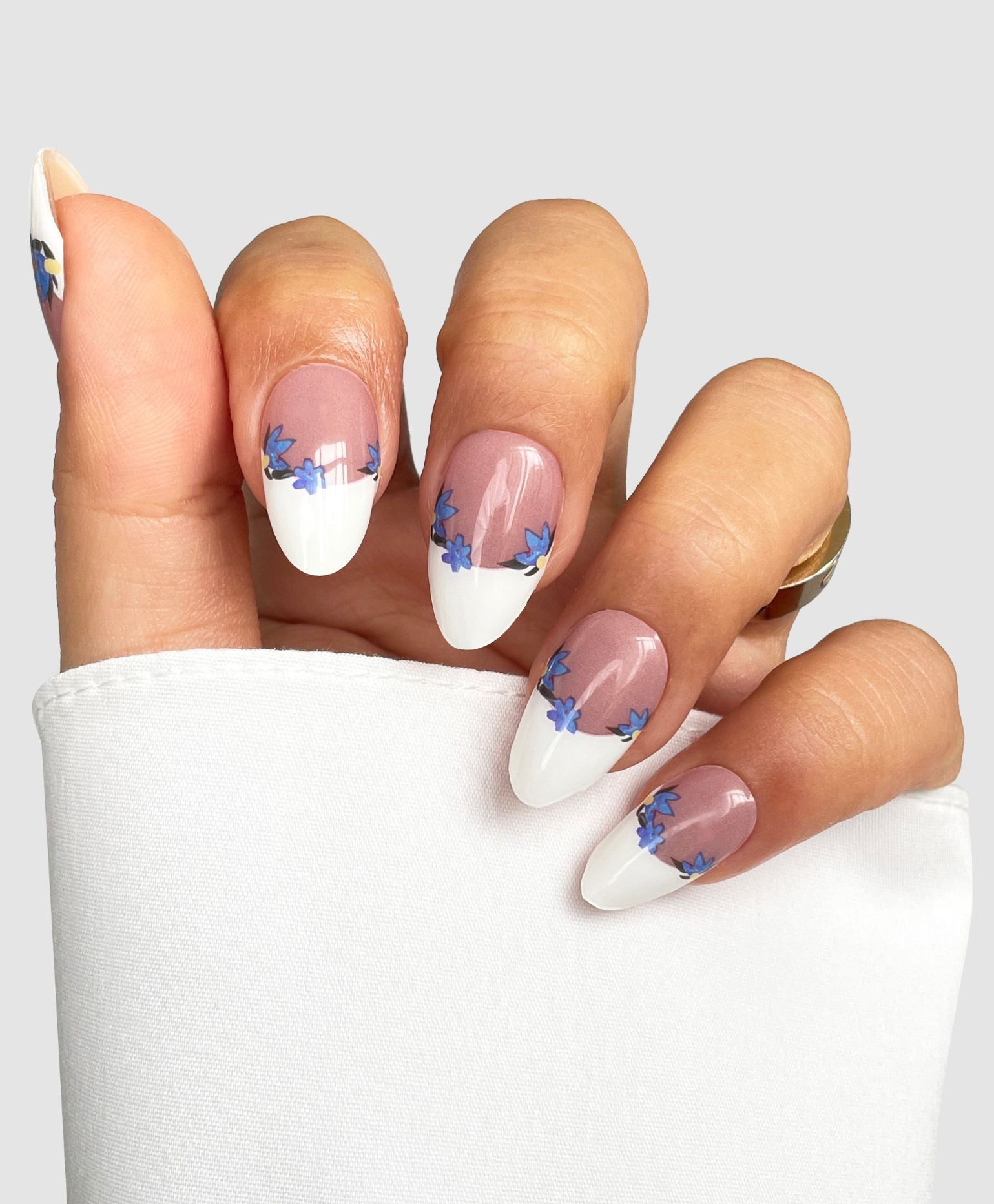 A hand with almond-shaped nails features Sassy Saints' French Bloom Gel Wraps—white French tip manicure with blue flowers—against a light background, complemented by a white long-sleeve top and gold rings.