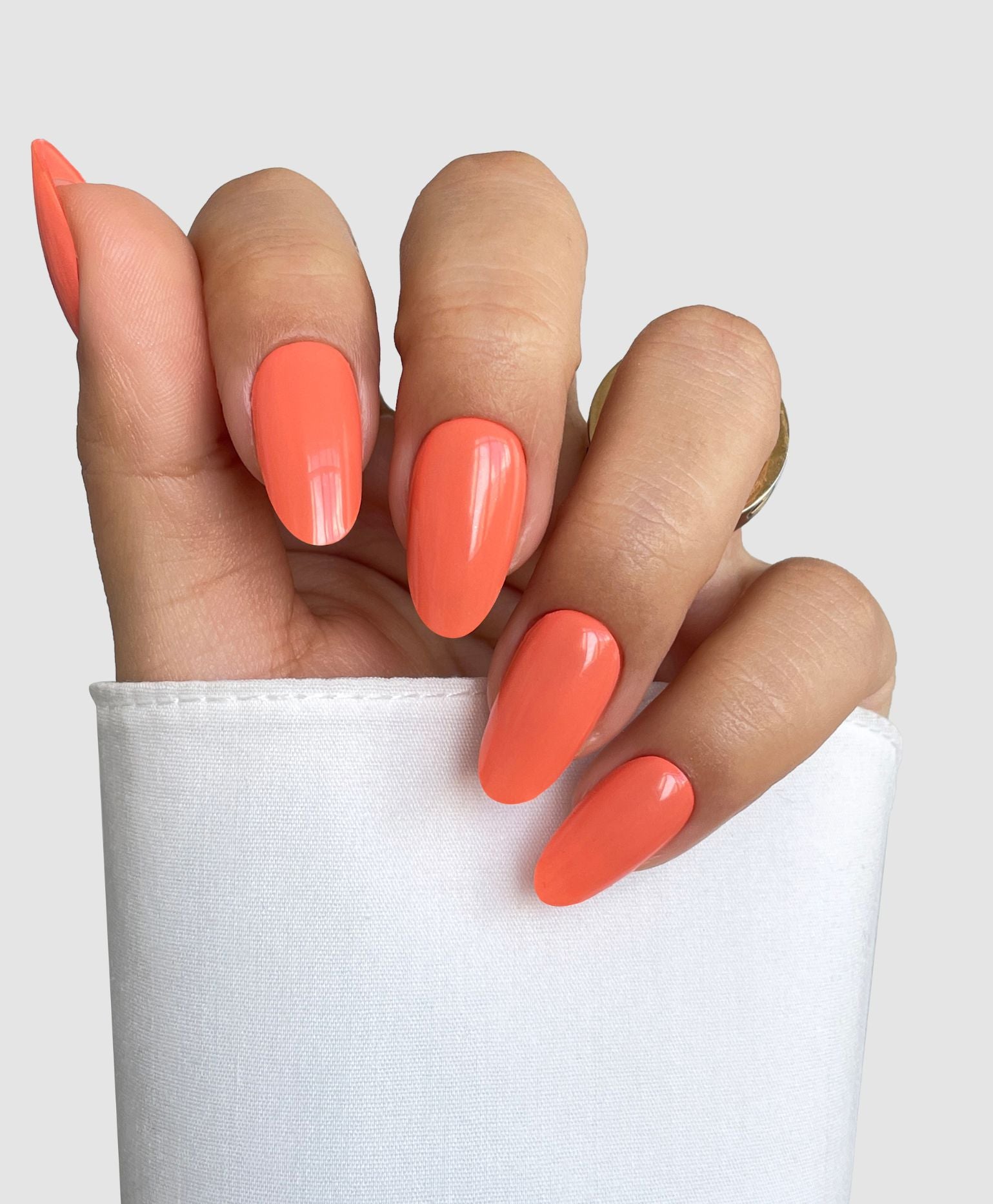 A hand with long, almond-shaped nails shows off Sassy Saints' Papaya Glow Gel Wrap in a glossy coral-orange, holding a white cloth against a light gray background.