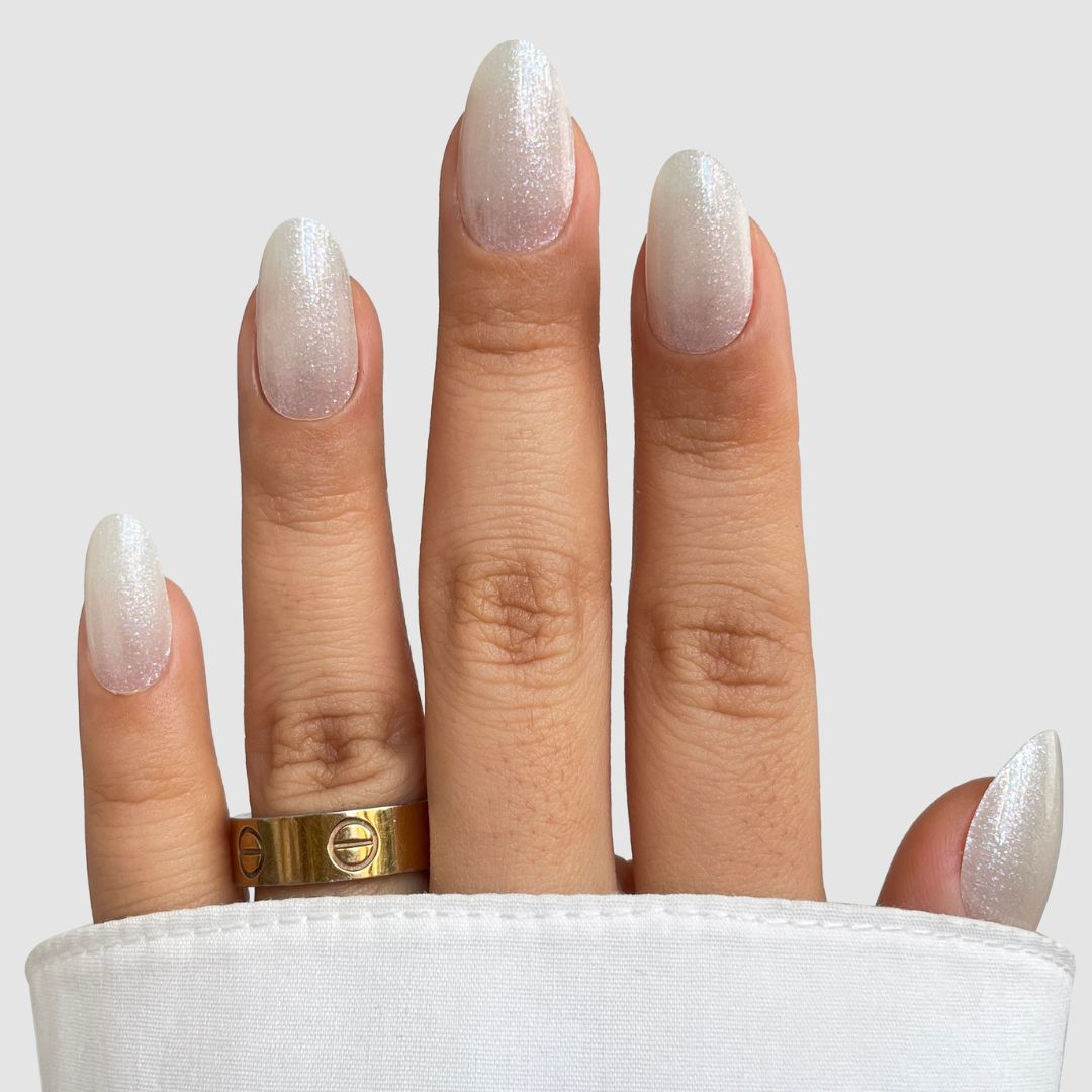 A hand with almond-shaped nails features Sassy Saints' Icy Veil Gel Wrap, a shimmery white ombre finish. Two gold rings adorn the middle finger, while a white fabric partially covers the hand.