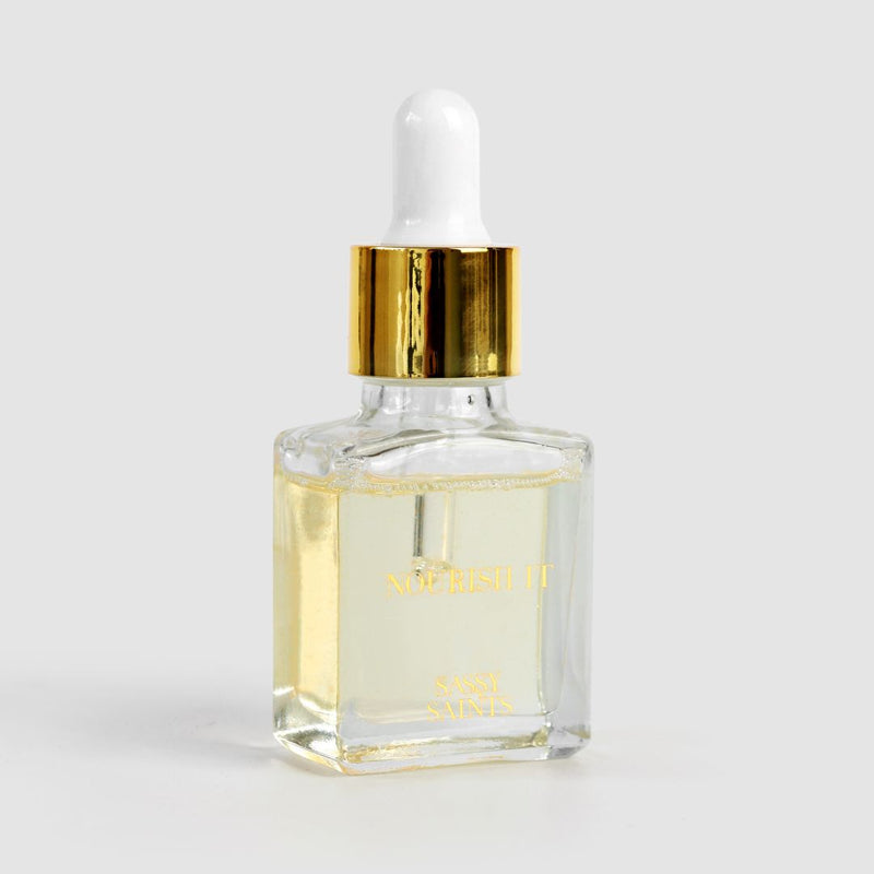 A clear glass dropper bottle with a gold and white cap holds light yellow Sassy Saints Cuticle Oil, set against a plain light gray background—the perfect nail care solution from Sassy Saints - Brings The Salon Home.