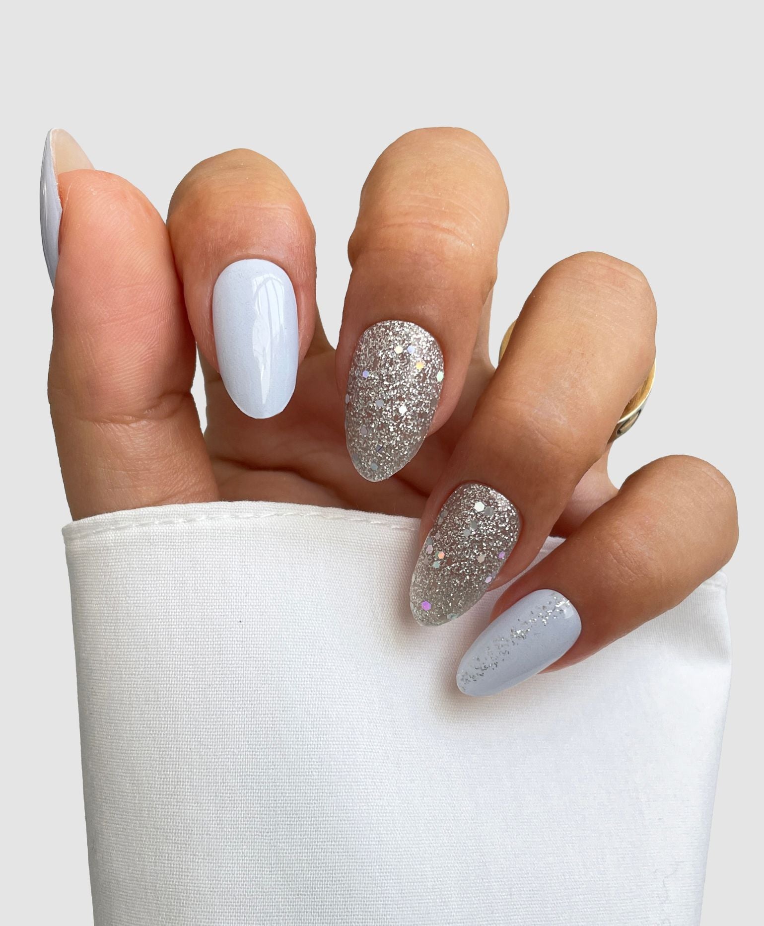 A hand with almond-shaped nails wears Sassy Saints' Hazy Frost Gel Wraps—light blue polish with silver glitter accents—partially covered by a white sleeve, set against a plain light background.