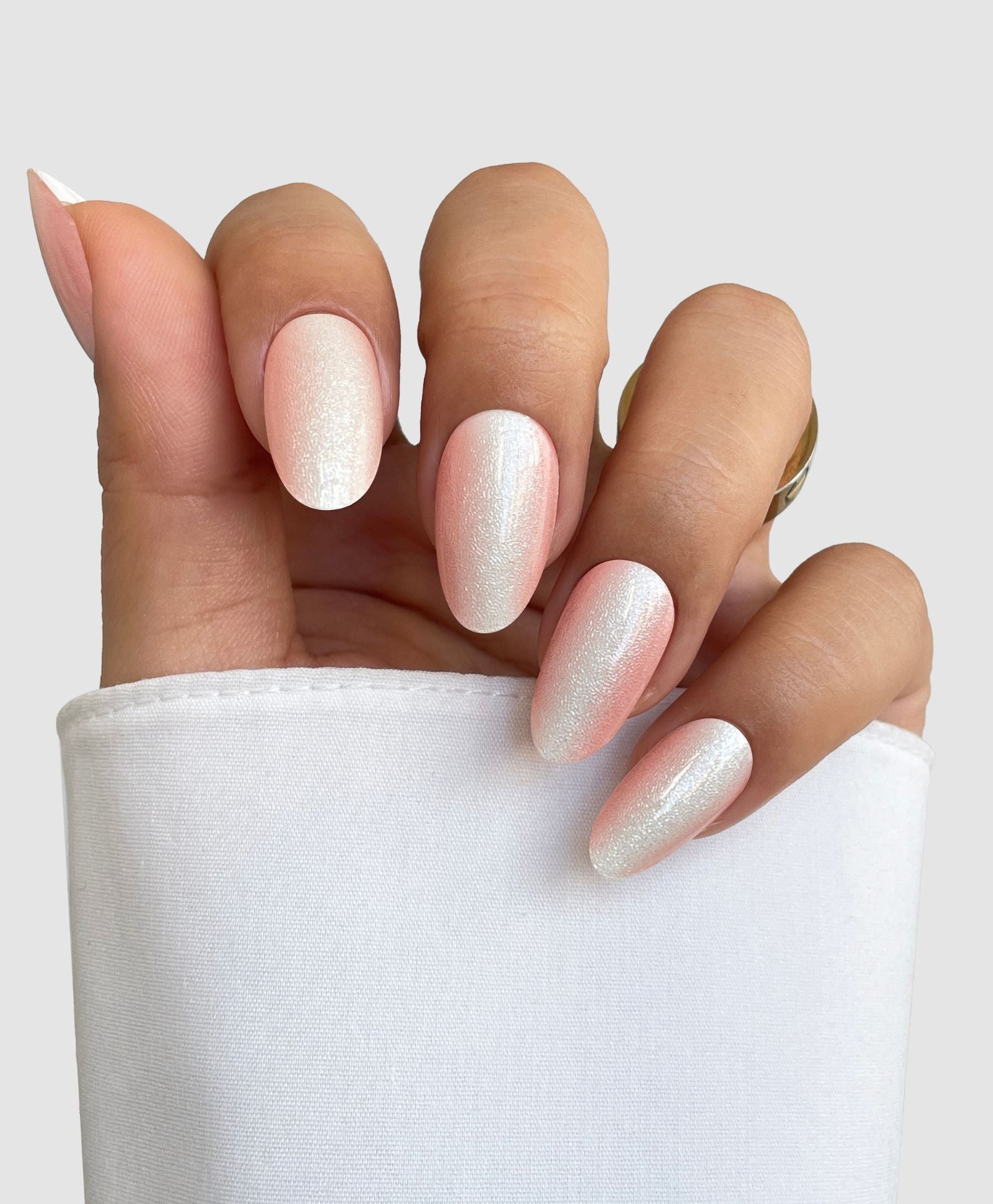 A hand with almond-shaped nails painted in Sassy Saints - Brings The Salon Home Silk Finish Gel Wrap, a shimmery pale pink, is shown against a plain light background. The person wears a white long-sleeve and a gold ring.
