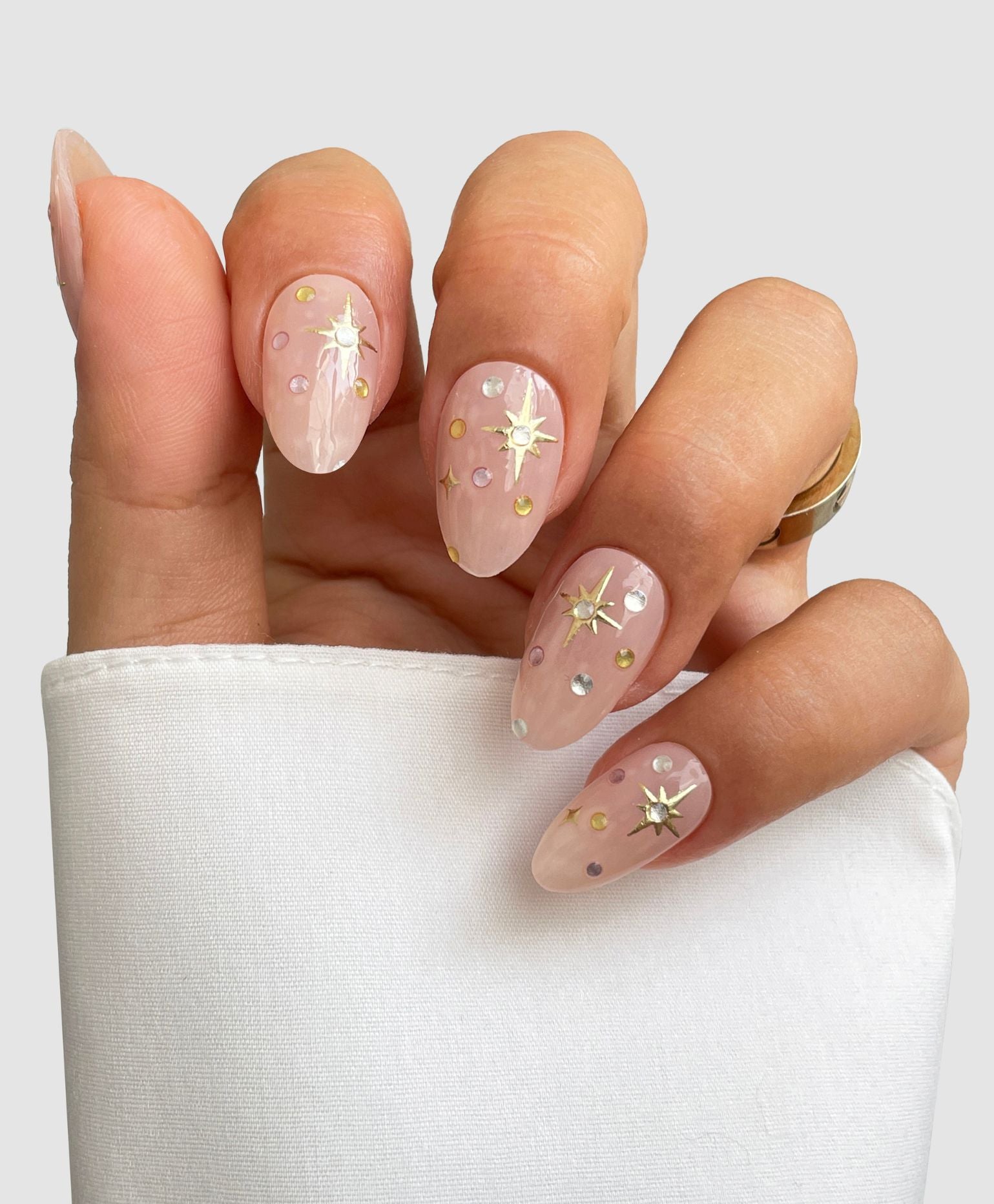 A hand showcases Sassy Saints' Milkyway Gel Wrap—almond-shaped, light pink nails with gold, silver, and iridescent star and dot accents—partially covered by a white sleeve against a plain background.