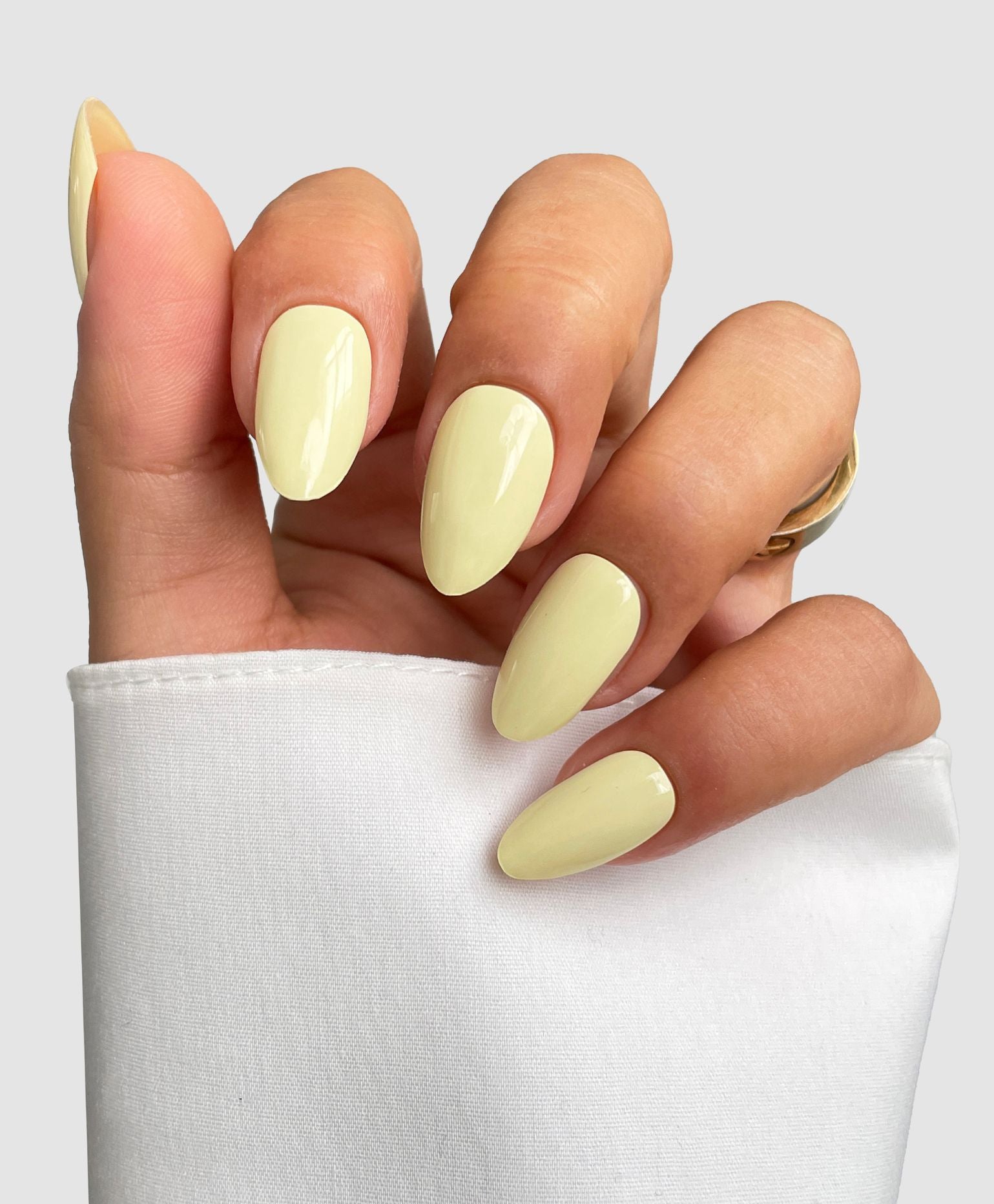 A hand with long, almond-shaped nails showcases Sassy Saints' Buttercup Yellow Gel Wraps against a plain backdrop, paired with a white sleeve and gold ring. Brings The Salon Home.
