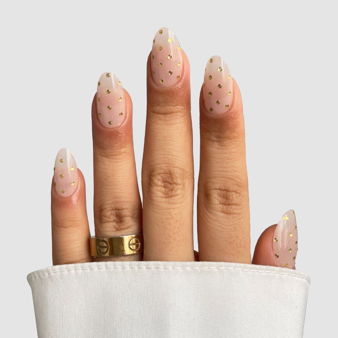 A hand showcases Sassy Saints - Brings The Salon Home's Champagne Drops Gel Wrap, featuring almond-shaped nails in sheer nude with gold dots, paired with a gold ring and a white sleeve against a plain light gray background.