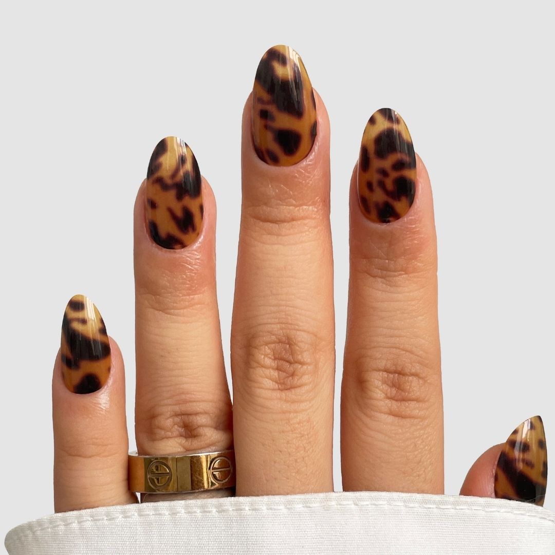 A hand with almond-shaped nails wears Sassy Saints' Shell Luxe Gel Wrap in glossy brown and black tortoiseshell, paired with two gold rings and shown on a light background featuring white fabric at the bottom.