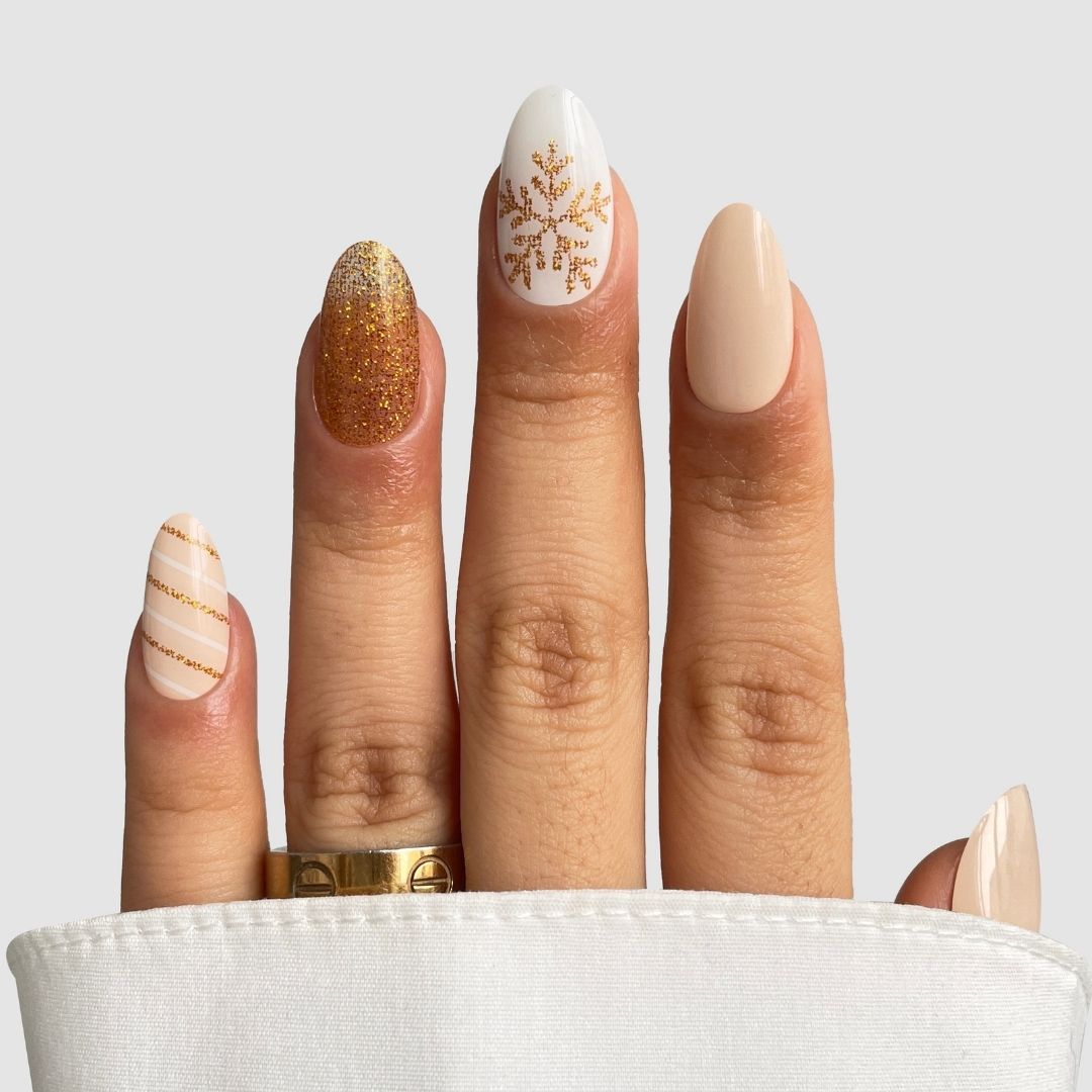 A hand with almond-shaped nails wears Sassy Saints - Brings The Salon Home’s Golden Flurry Gel Wrap: festive gold glitter, gold snowflake on white, beige, and striped accents. A white sleeve and a gold ring complete the look.