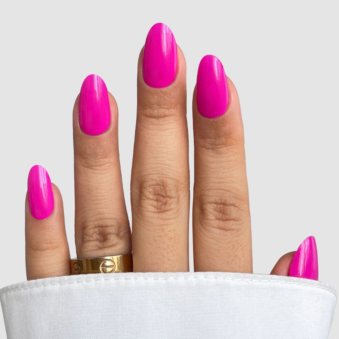 A hand with glossy, almond-shaped nails enhanced by Pink Hype Gel Wrap from Sassy Saints – Brings The Salon Home, rests on a white surface. A gold ring adorns the middle finger and the background is light gray.