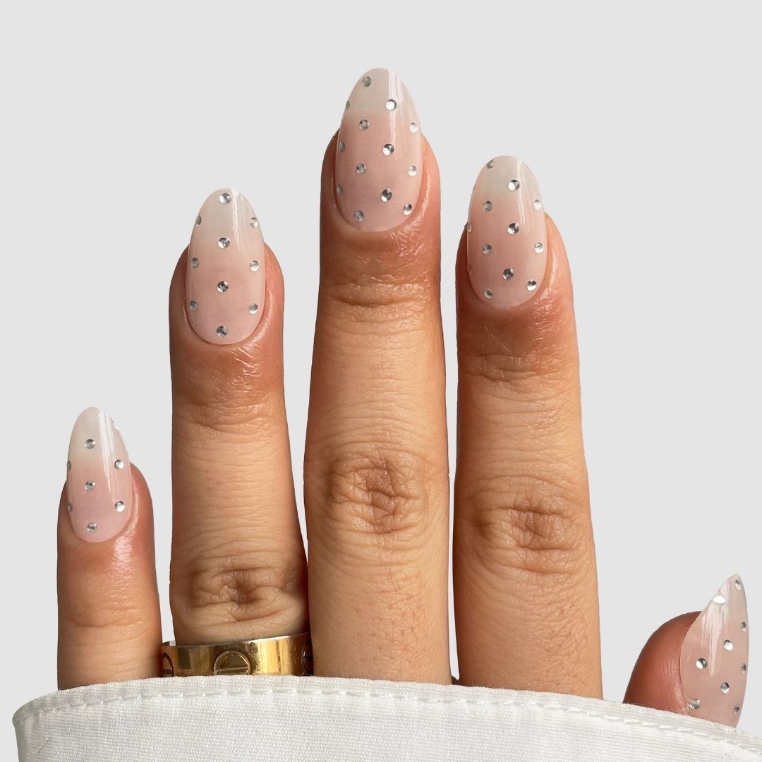 A hand shows off Sassy Saints’ Platinum Spotlight Gel Wrap—sheer nude-pink with small silver rhinestones on each almond nail. A gold ring and the cuff of a white shirt complete the look.