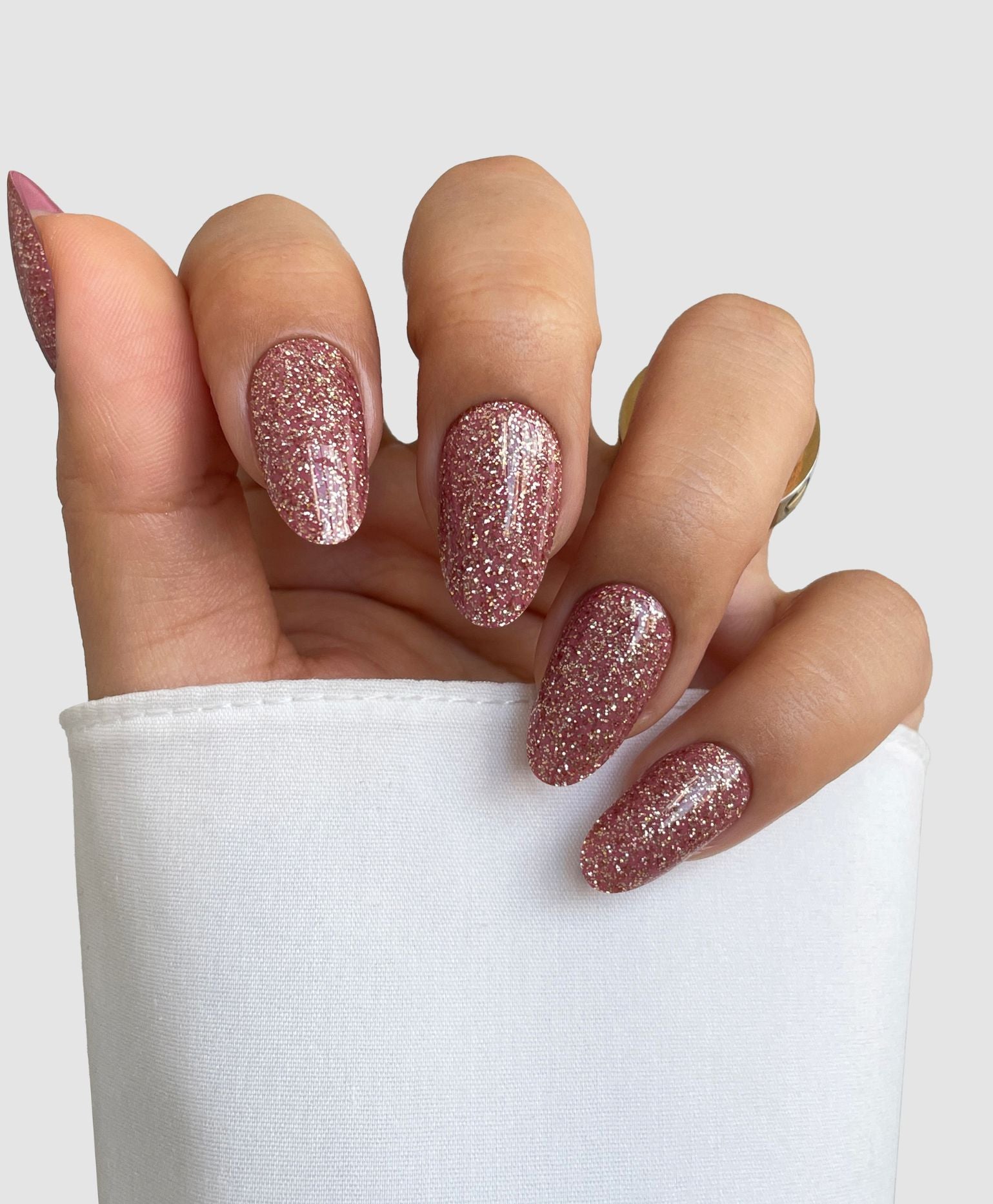 A hand with almond-shaped nails features Sassy Saints' Rose Spark Gel Wrap, a sparkly pink glitter look, against a plain light grey background. The person wears a white sleeve and a gold ring.