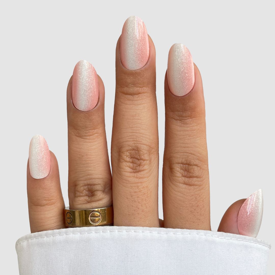 A hand with Silk Finish Gel Wrap by Sassy Saints - Brings The Salon Home, shows neatly manicured pink and white ombre nails, two gold rings on the middle finger, partially covered by a white sleeve, set against a light grey background.