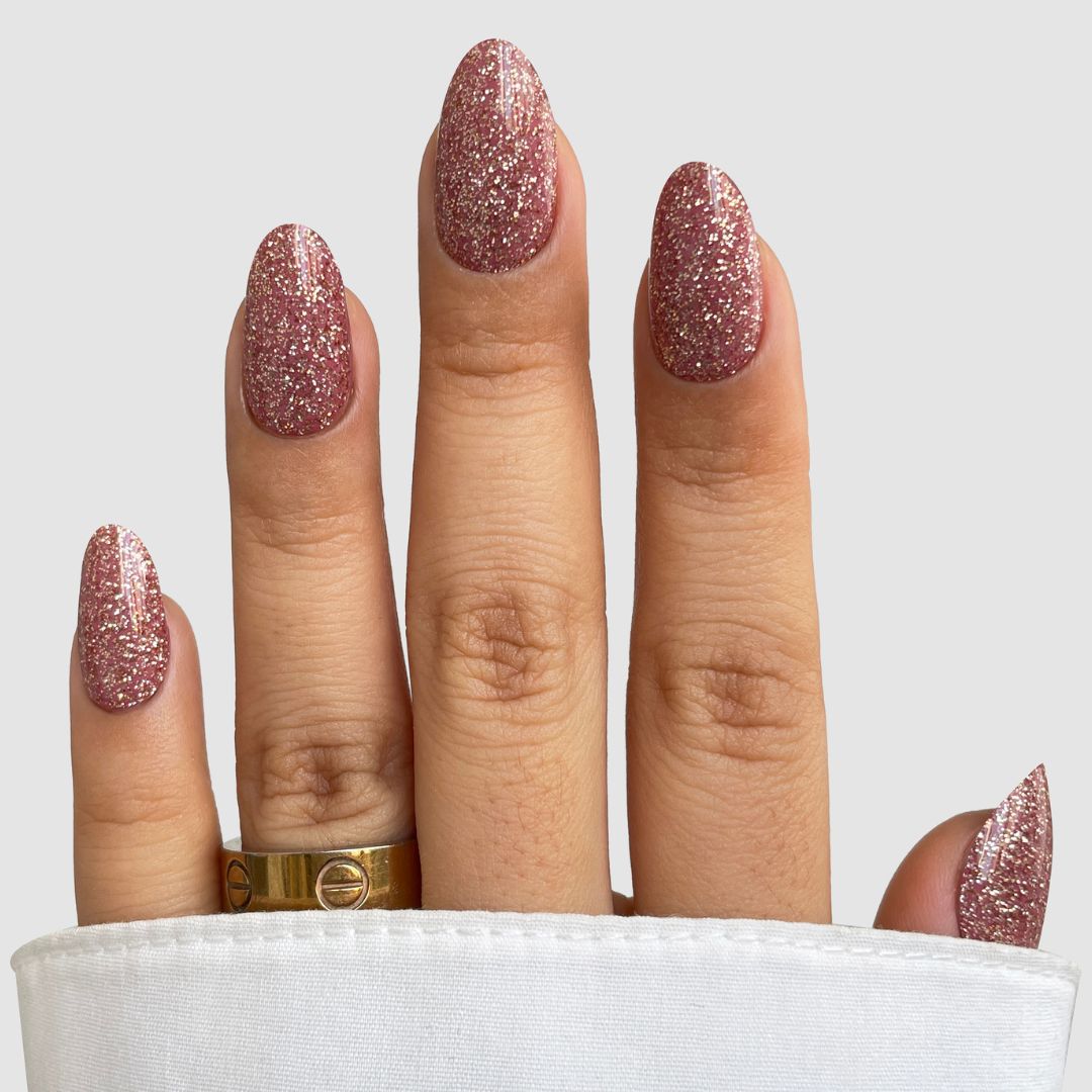 A hand wears Sassy Saints' Rose Spark Gel Wrap, a pink glittery nail wrap, with a gold ring on the ring finger and a white sleeve, set against a plain light background.