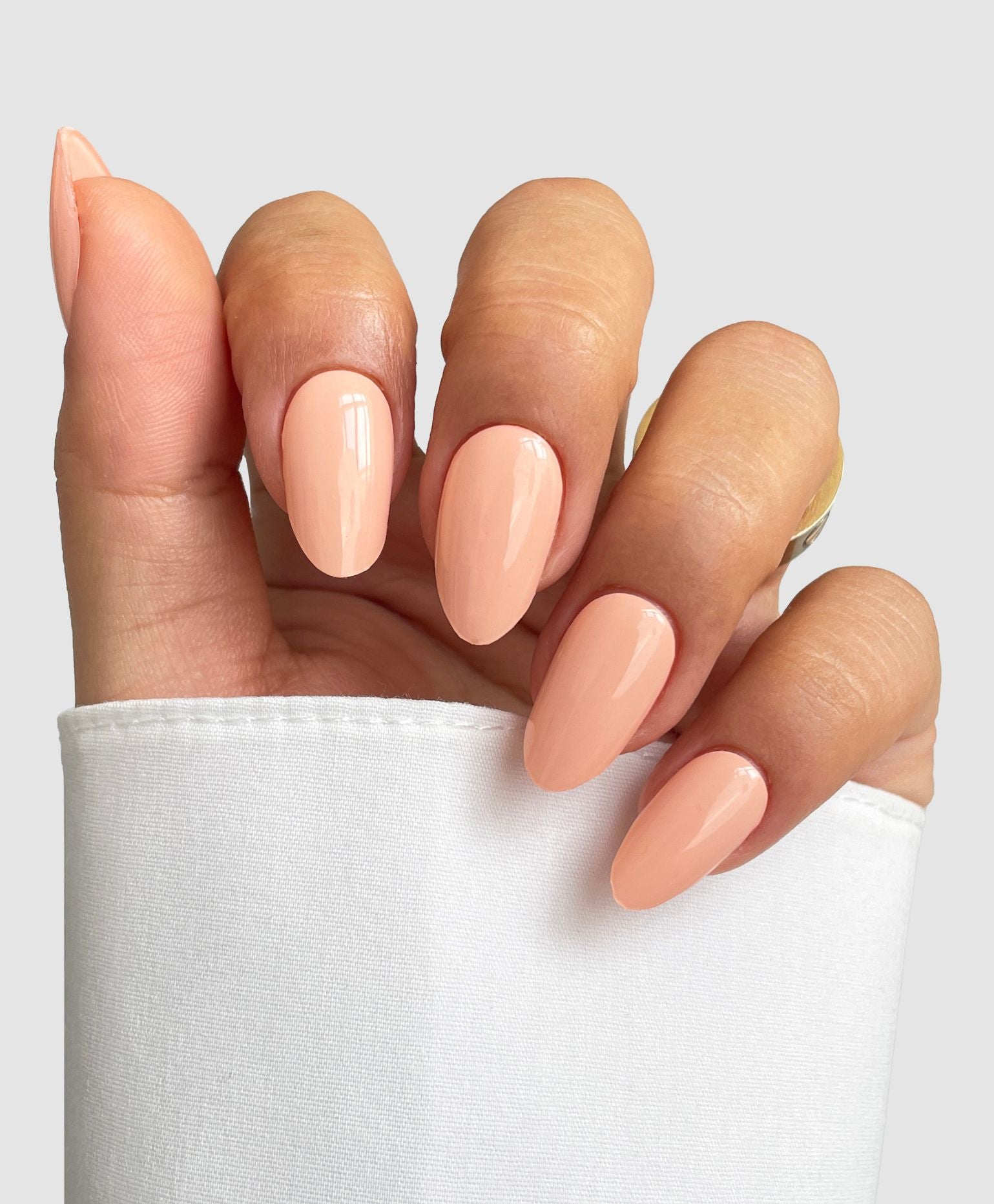 A hand with long, almond-shaped nails features Sassy Saints' Apricot Mood Gel Wrap in a glossy peach shade, partially covered by a white sleeve and set against a plain light gray background.