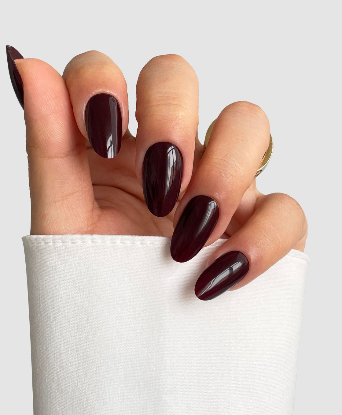 A hand with long, almond-shaped nails painted in Sassy Saints' Dark Current (Dark Red) rests against a white shirt sleeve, with gold rings on two fingers.