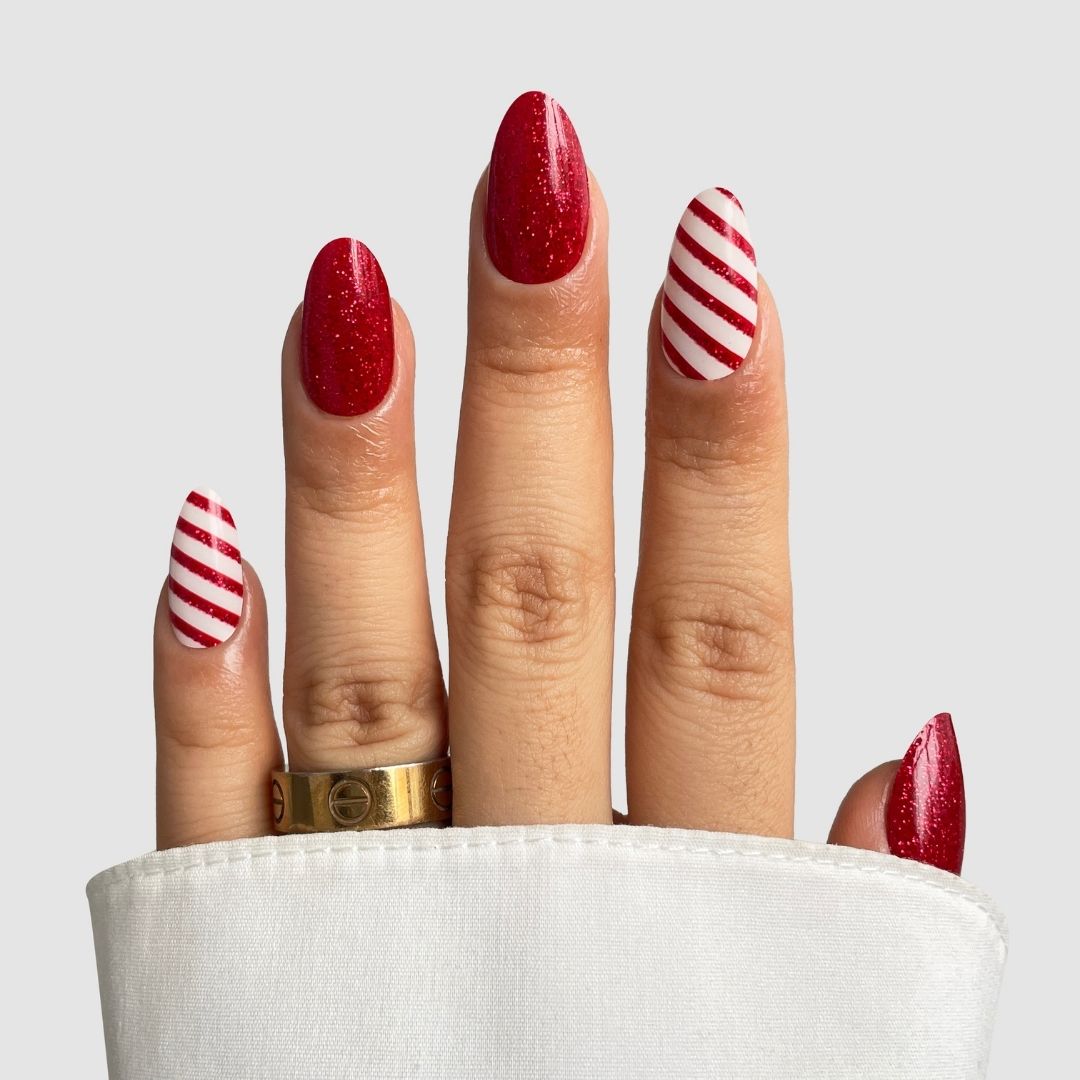A hand sports Sassy Saints' Candy Cane Gel Wraps—almond-shaped red nails with two featuring diagonal white and red candy cane stripes. A gold ring and white sleeve complete the look against a plain light background.