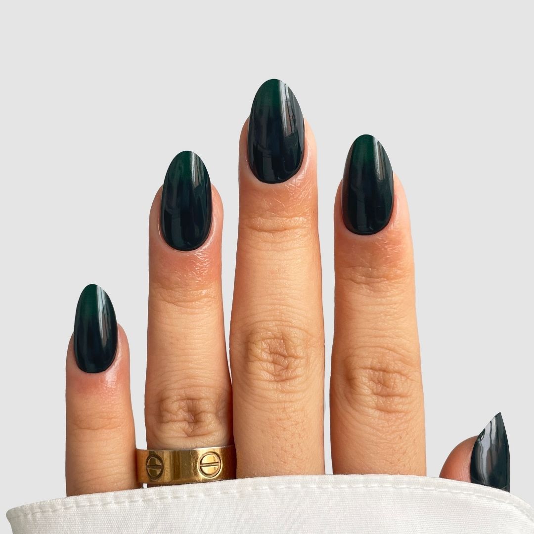 A hand with almond-shaped nails painted in Sassy Saints’ Velvet Fir (Dark Green) polish, featuring a gold ring on the middle finger and partially covered by white fabric, set against a light gray background.