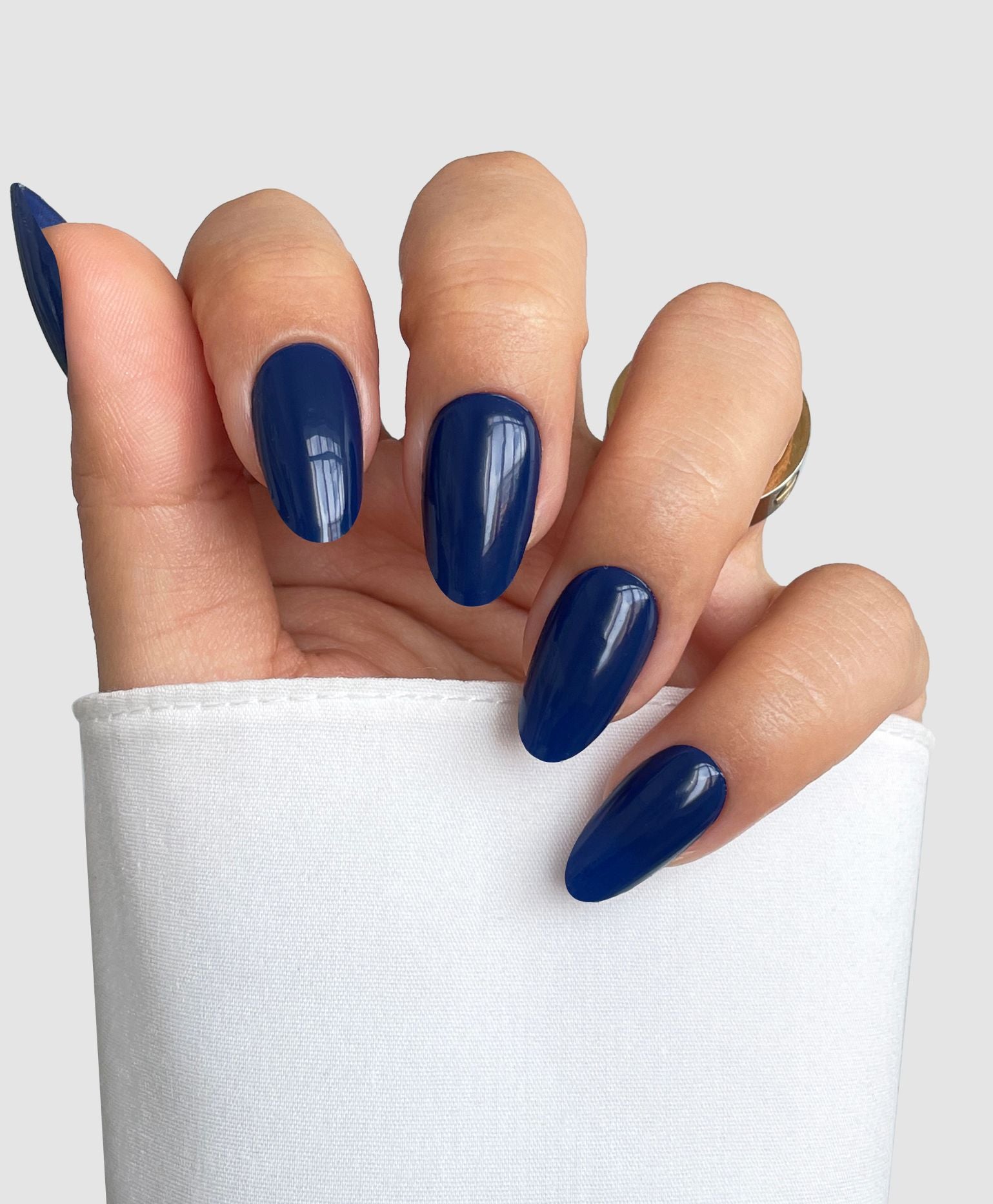 A hand with long, oval nails wears Sassy Saints - Brings The Salon Home's Blue Eclipse Gel Wrap in glossy navy blue, resting on white fabric. A gold ring decorates the middle finger against a neutral background.