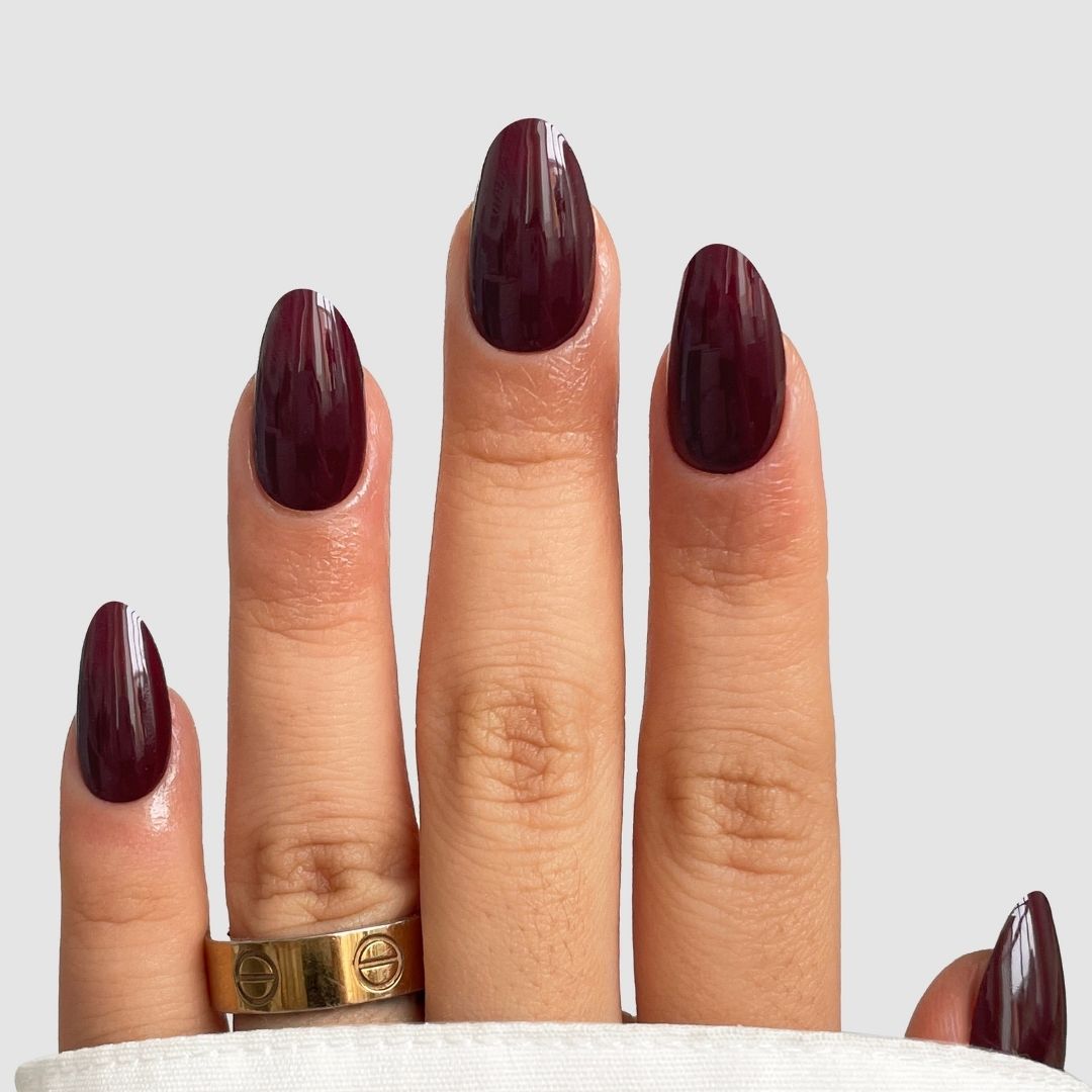 A hand with long, almond-shaped nails in Sassy Saints' Dark Current (Dark Red), a glossy maroon shade, wearing a gold ring on the middle finger against a plain light gray background.