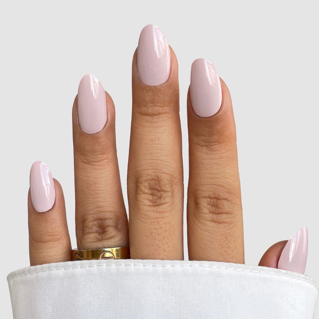 A hand with almond-shaped nails in glossy pale pink Soft Tulle Gel Wrap by Sassy Saints - Brings The Salon Home, wearing a gold ring on the middle finger and partially covered by white fabric.