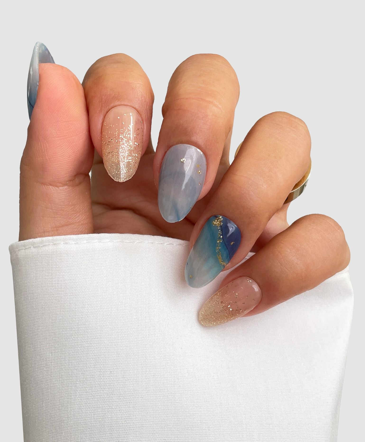 A hand with almond-shaped nails features Sassy Saints' Ocean Dream Gel Wrap, showcasing nude glitter and blue marble designs, partially covered by a white sleeve against a light grey background.