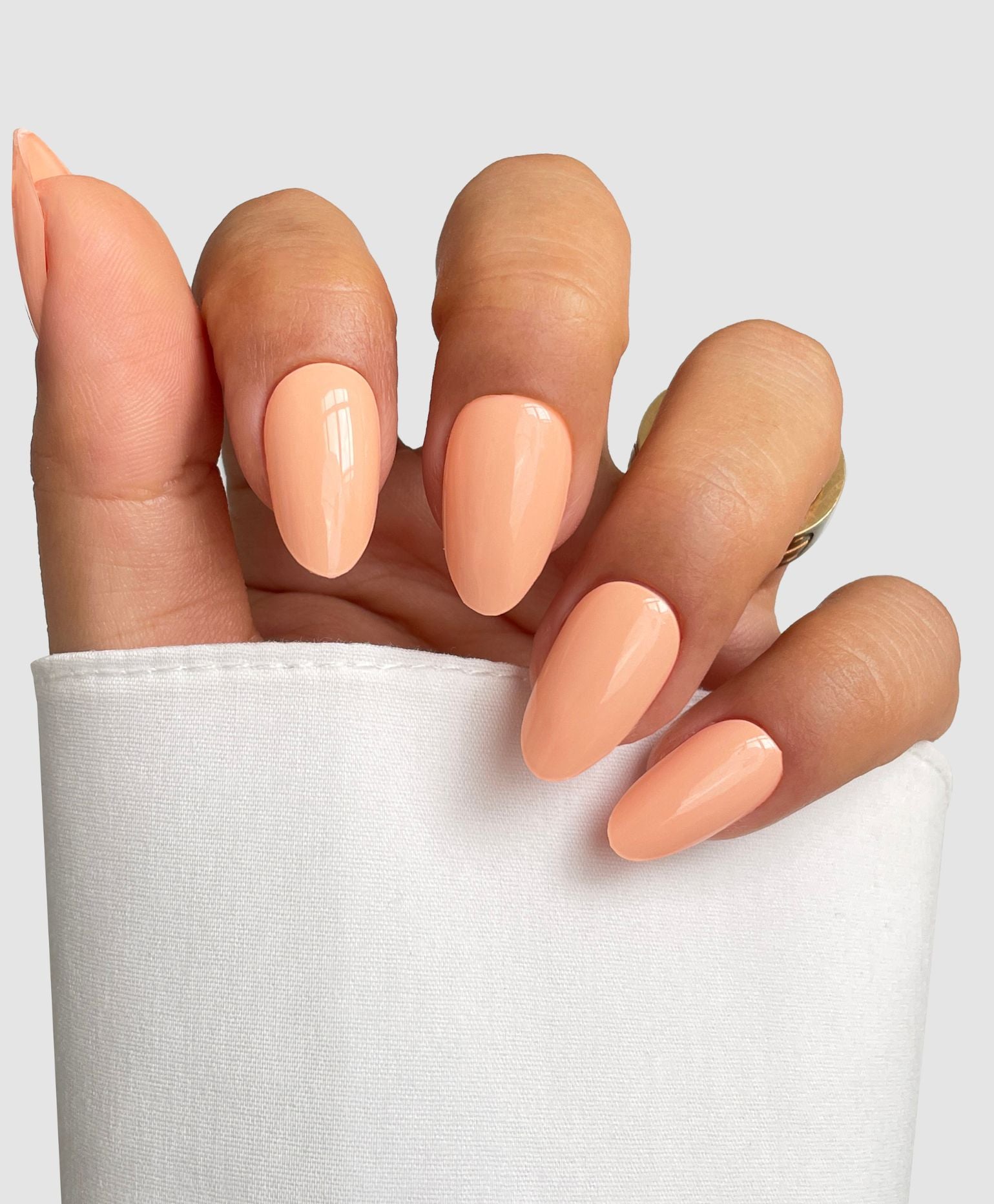 A hand with almond-shaped nails in Sassy Saints’ Peach Glow Gel Wrap rests against a light gray background. The glossy peach nails, paired with a gold ring and a white sleeve, showcase salon-quality style at home.