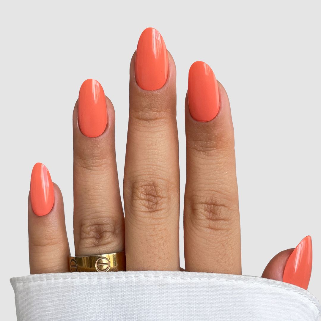 A hand with medium-length almond-shaped nails painted in Sassy Saints' Papaya Glow Gel Wrap, wearing a gold ring and partially covered by white fabric, is set against a plain light gray background.