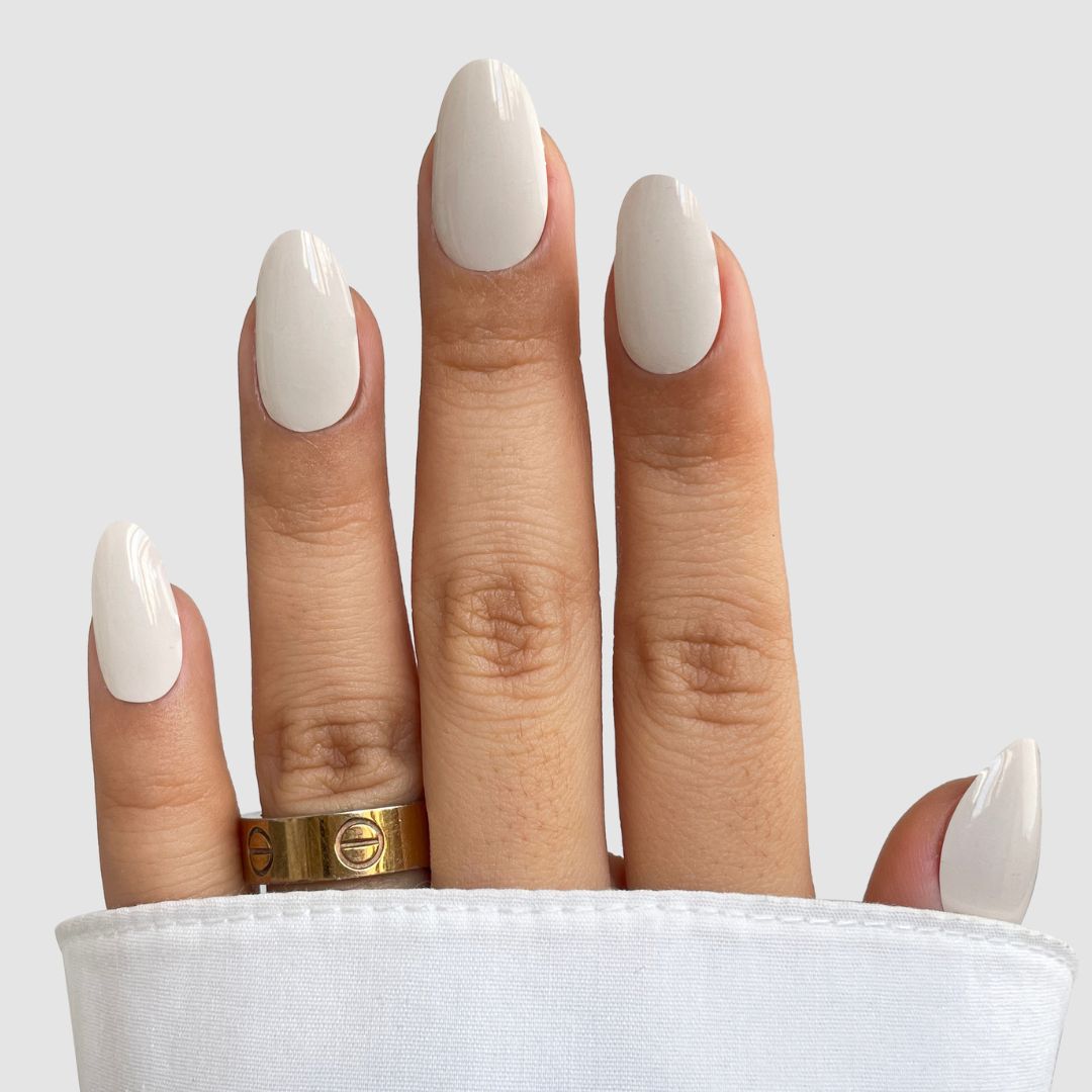 A hand with long oval nails in glossy light beige Blush Milk Gel Wrap by Sassy Saints - Brings The Salon Home, wearing a gold ring on the middle finger and partially covered by a white sleeve against a plain background.
