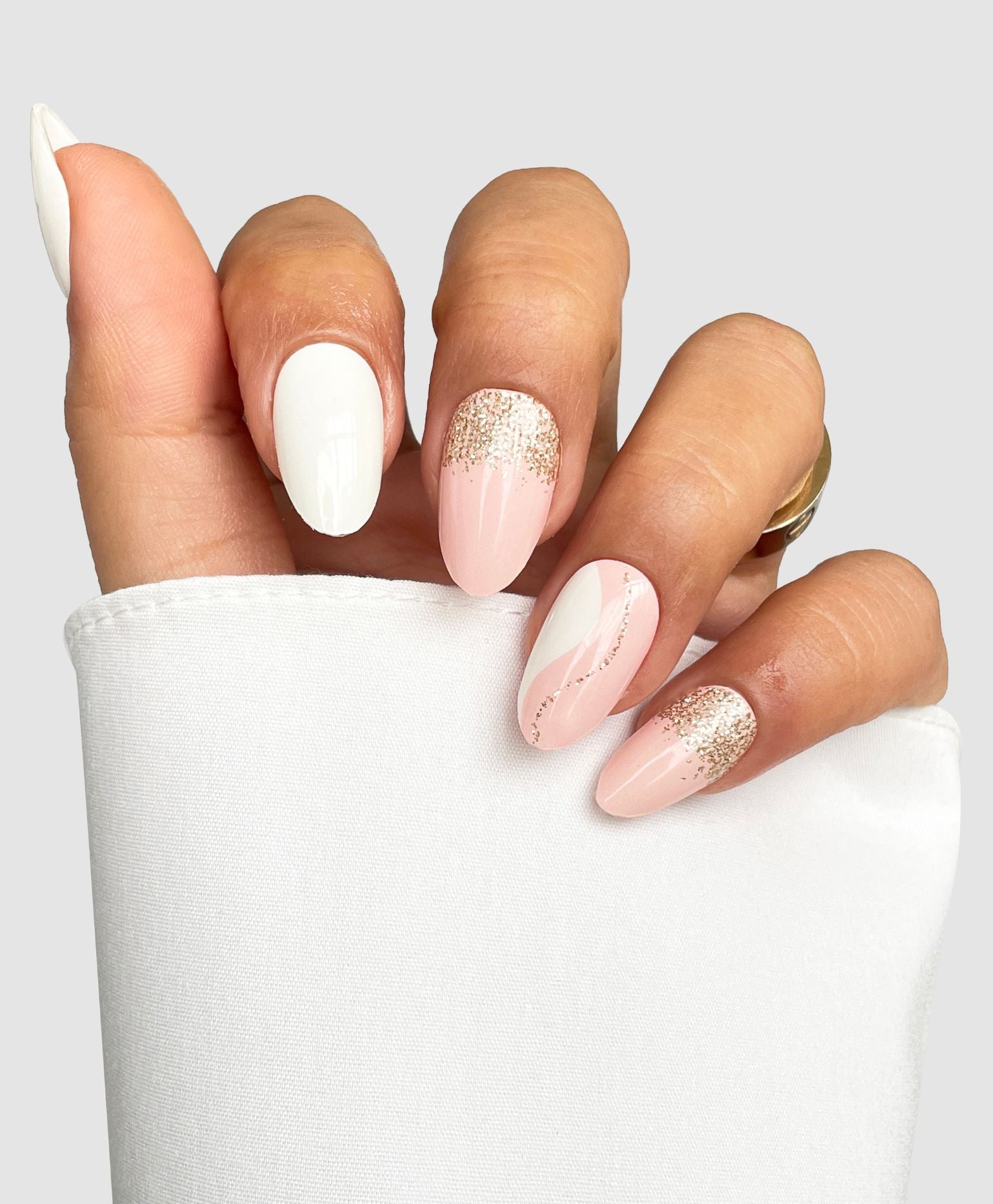 A hand with almond-shaped nails shows off Sassy Saints - Brings The Salon Home’s Soft Launch Gel Wrap in a white, nude pink, and glittery gold design while holding a white fabric.