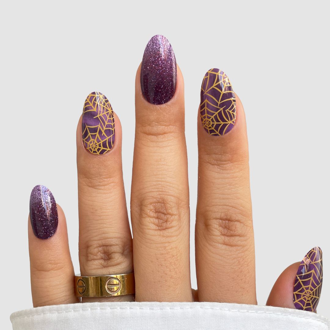 A hand displays Sassy Saints - Brings The Salon Home's Wicked Web Gel Wrap: purple glittery nails with gold spiderwebs, accessorized with two gold rings, set against a plain light background.