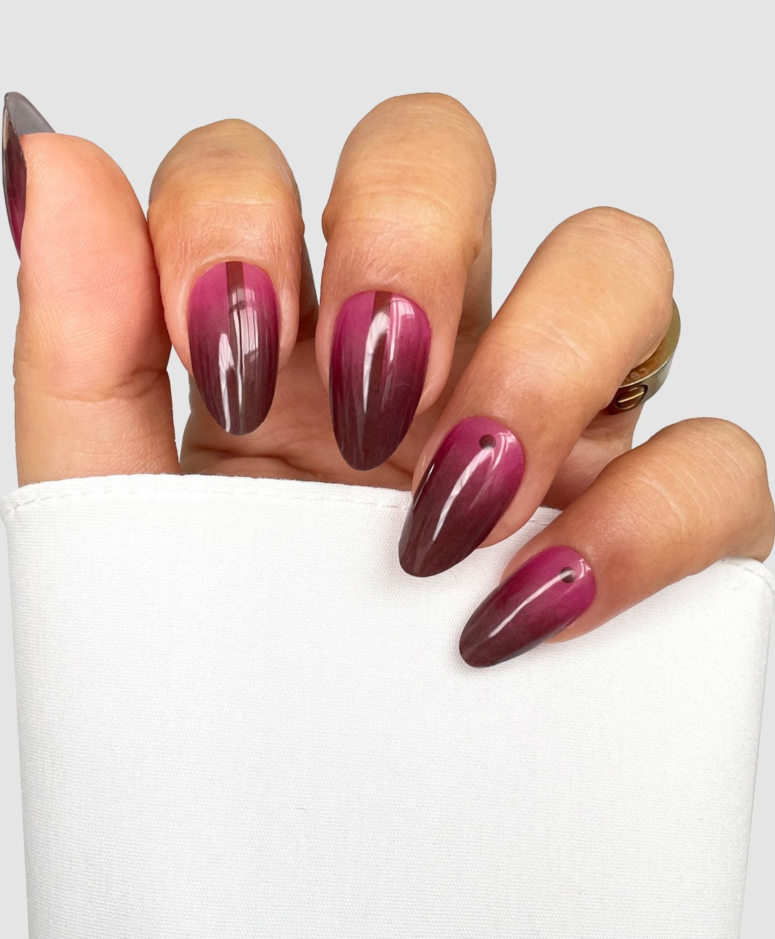 A hand with long, almond-shaped nails featuring Sassy Saints' Cherry Smoke Gel Wrap in glossy dark red and pink tones holds a white cloth against a light gray background.