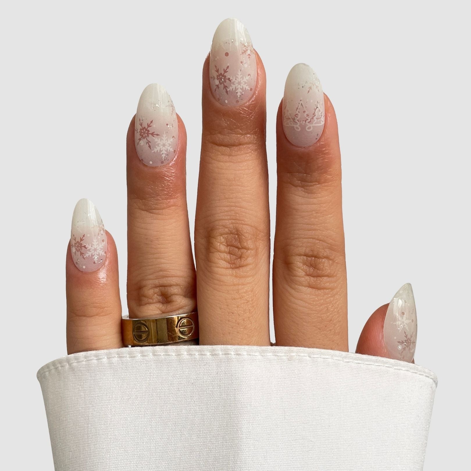 A hand shows Sassy Saints - Brings The Salon Home’s Arctic Frost Gel Wrap: almond-shaped nails with white, snowflake winter art. A gold ring adorns the ring finger, peeking from a white sleeve against a plain light gray background.