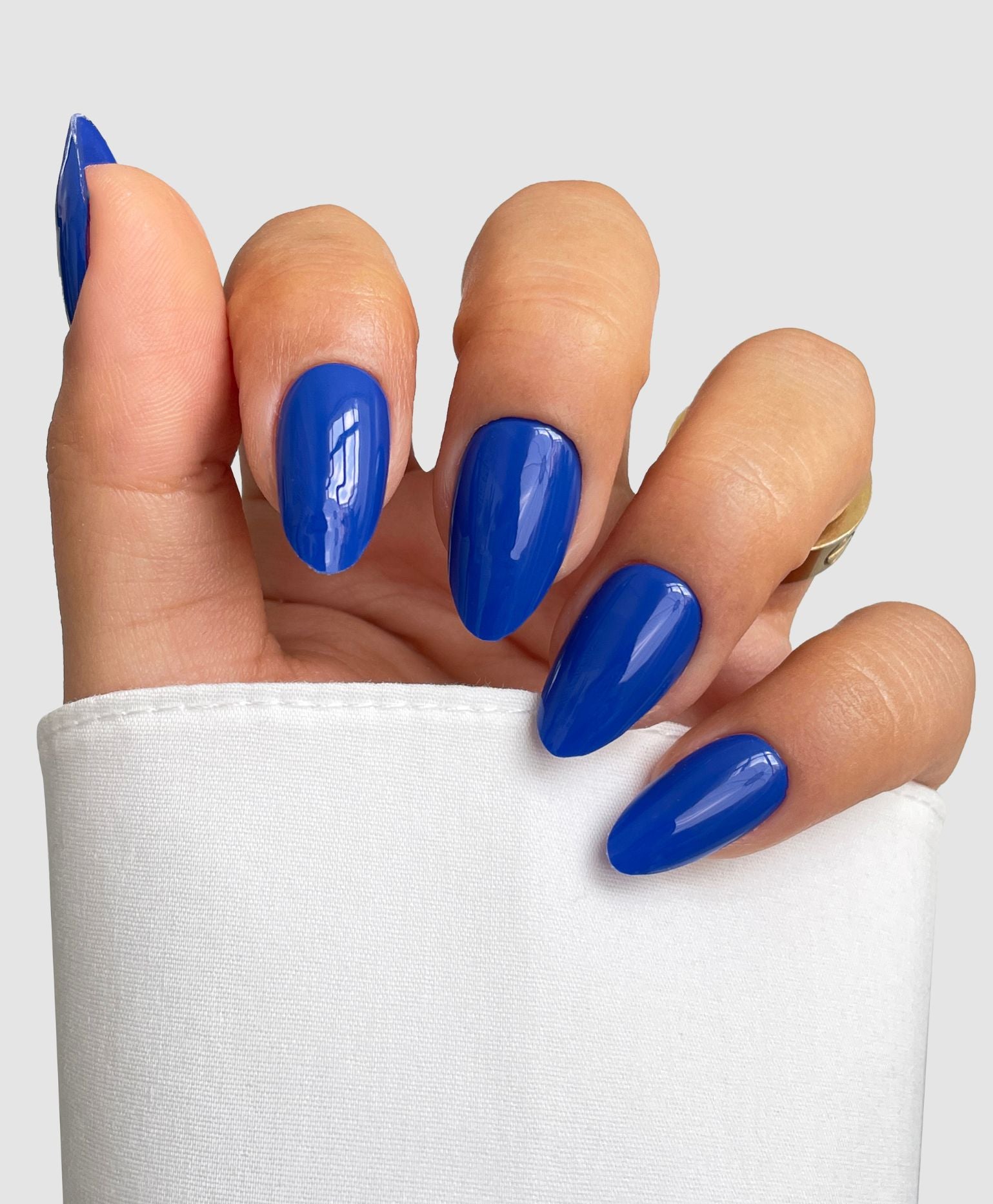 A hand with almond-shaped nails showcases Sassy Saints' Blue Me Away Gel Wrap, a shiny royal blue polish, partially covered by a white sleeve against a light gray background.