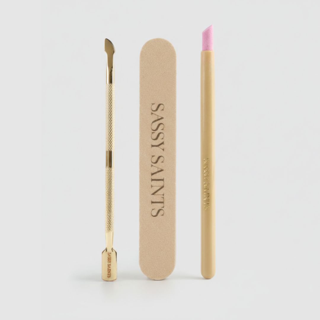 The Sassy Saints Perfect Prep Bundle (FREE)—featuring a gold cuticle pusher, a beige Sassy Saints nail file, and a gold cuticle oil pen with pink tip—is arranged vertically on a light gray background.