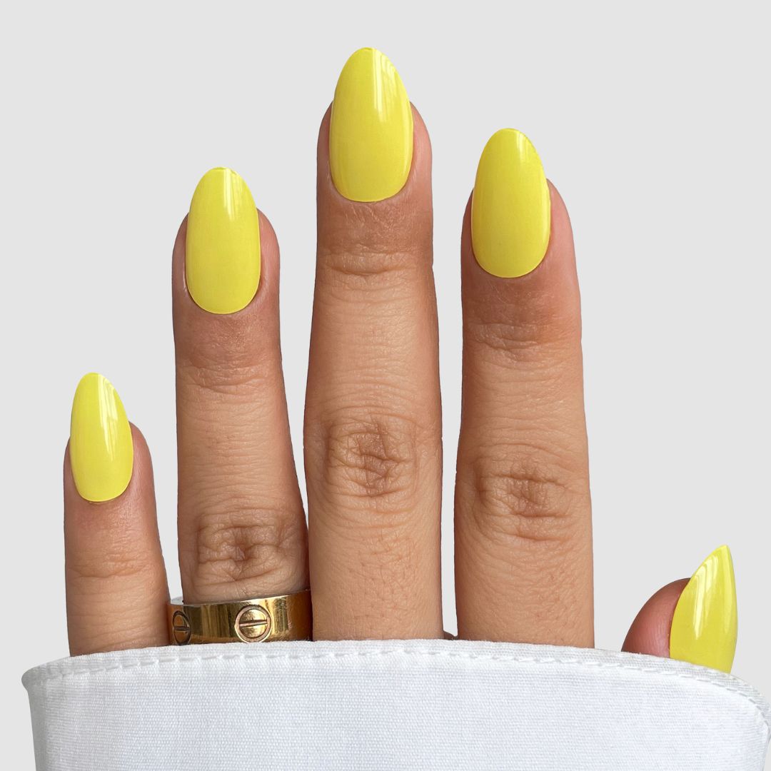 A hand with neatly manicured nails in Sassy Saints - Brings The Salon Home Citrus Pop Gel Wrap, wearing two gold rings and partially covered by white fabric, set against a plain light gray background.