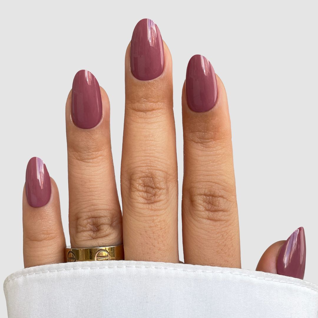 A hand with medium-length nails features Sassy Saints' Deep Petal Gel Wrap in glossy mauve, partially covered by a white fabric. The middle finger displays a gold engraved ring, set against a light gray background.