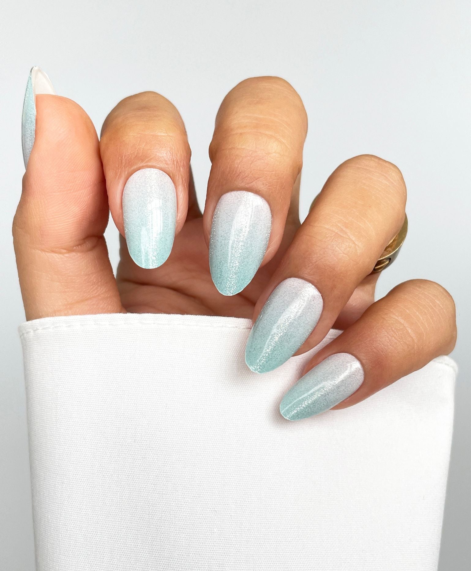 A hand with long, almond-shaped nails features Sassy Saints - Brings The Salon Home's Frozen Glow Gel Wrap in a pale blue to white shimmer gradient, posed against a white background with a gold ring and white sleeve.