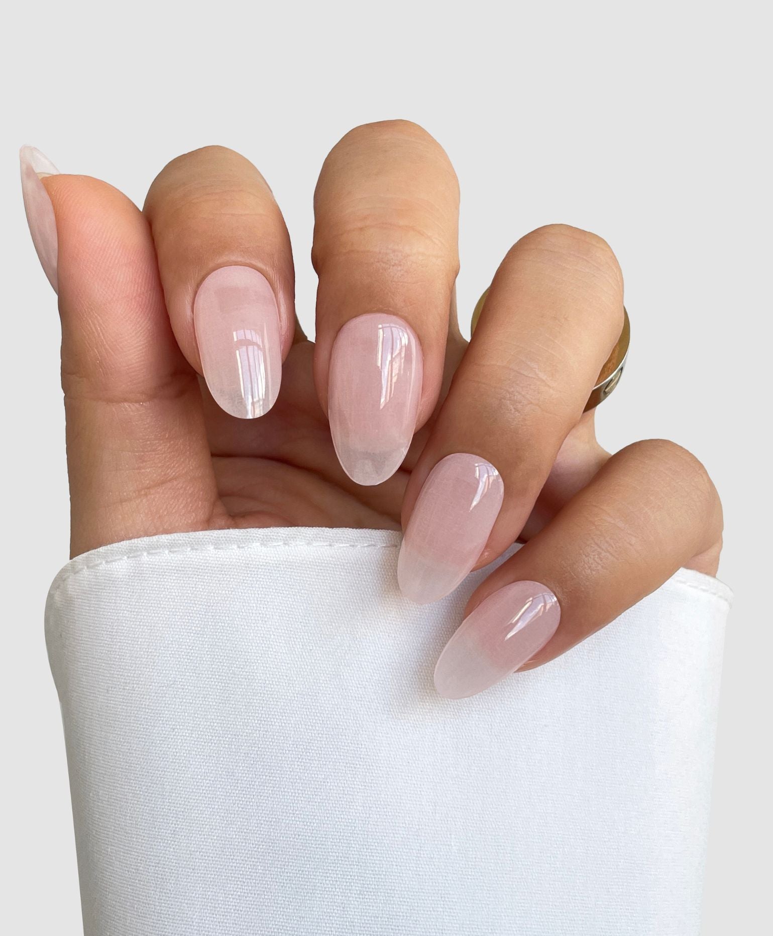 A hand with oval nails styled in Sassy Saints' Sugar Glaze Gel Wrap, a light glossy nude pink, partially draped in white fabric against a plain light gray background.