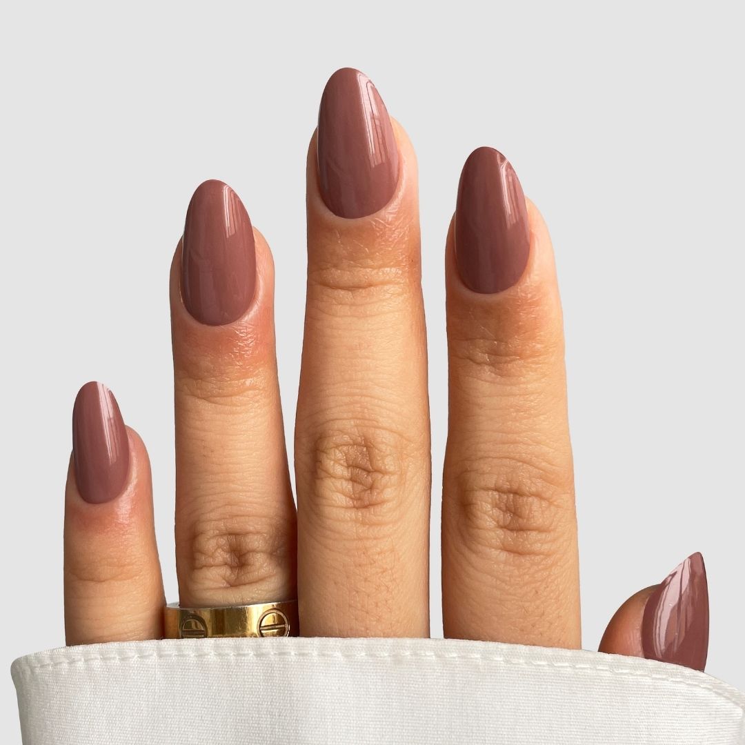 A hand with medium-length almond nails features Sassy Saints’ Baked Clay Gel Wrap, a glossy mauve polish. A gold ring adorns the middle finger, with white fabric partially covering the base of the fingers.