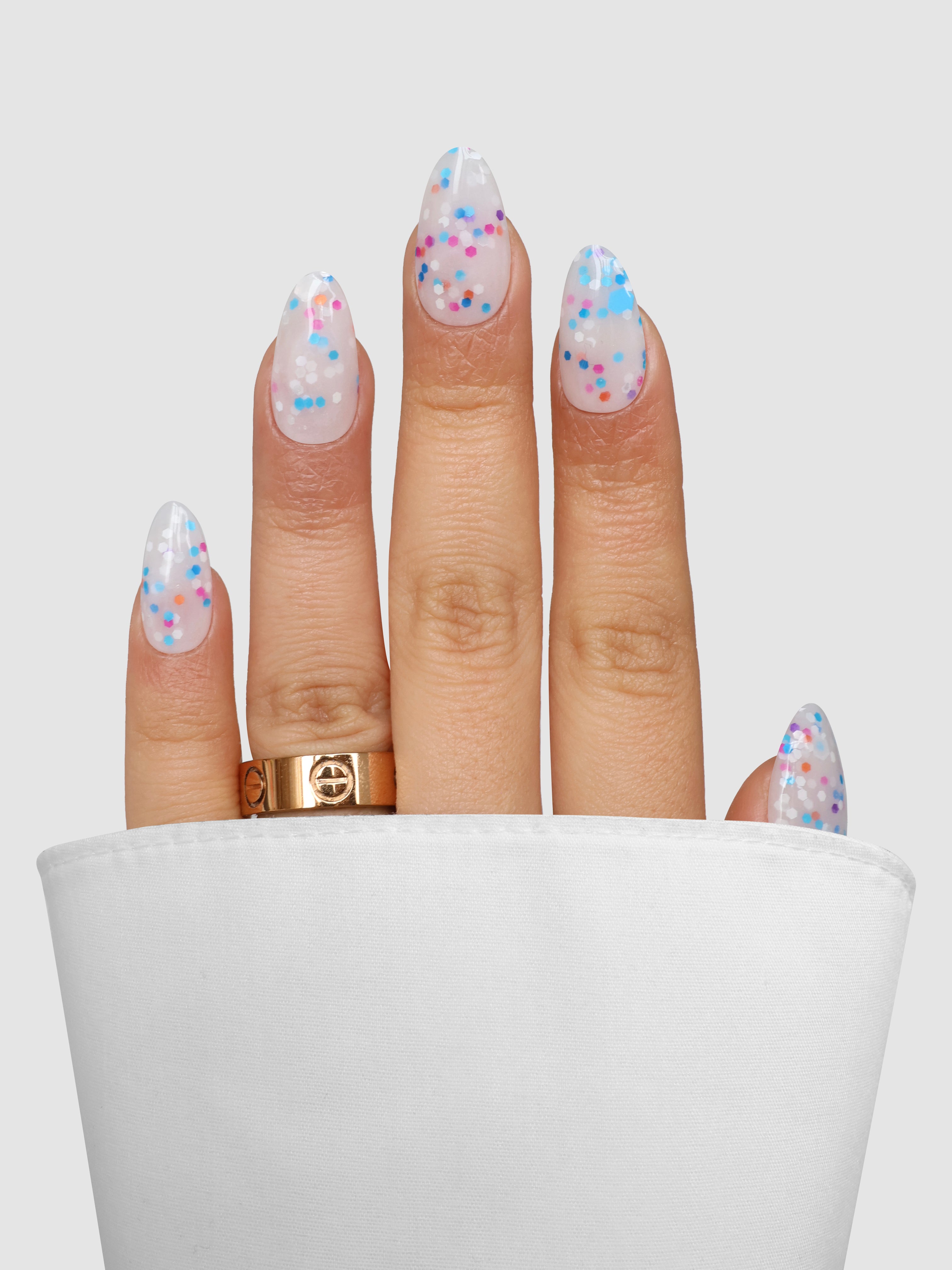A hand with almond-shaped nails coated in Sassy Saints' Sugar Pop Dipping Powder, showcasing a clear base with colorful confetti dots, wears a gold ring and is partially wrapped in white cloth against a light gray background.