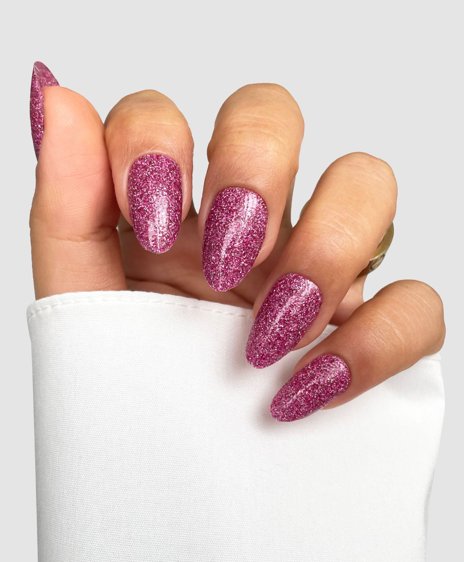 A hand with almond-shaped nails features Sassy Saints' Doll Moment Gel Wrap, a sparkly pink glitter polish, partially wrapped in a white sleeve against a plain light gray background.