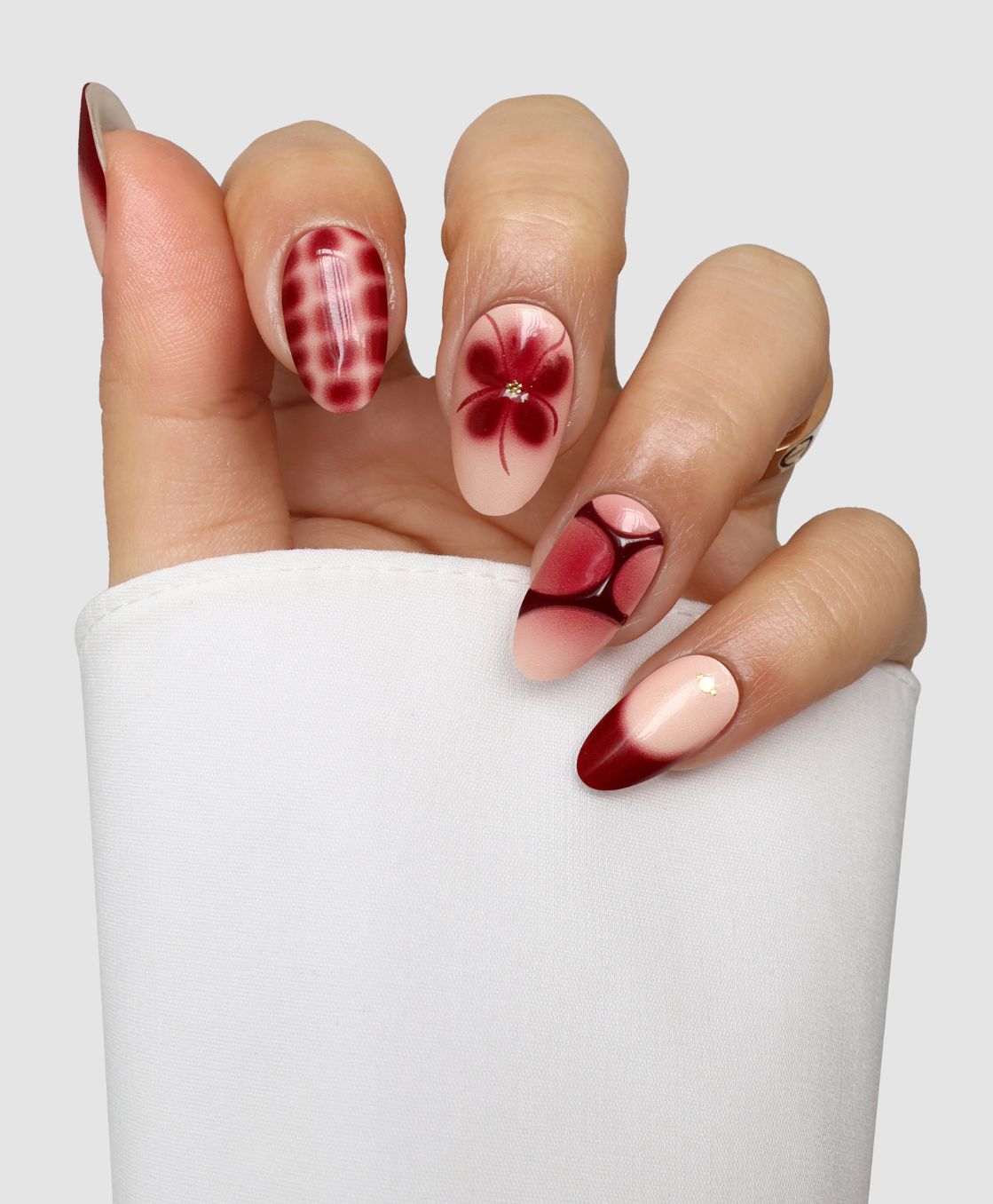 A hand displays Sassy Saints’ Burgundy Bloom (3D) Gel Wraps, showing red and nude gradient nails with plaid, gem-centered flower, geometric designs, and gold accents—all on a plain white background.