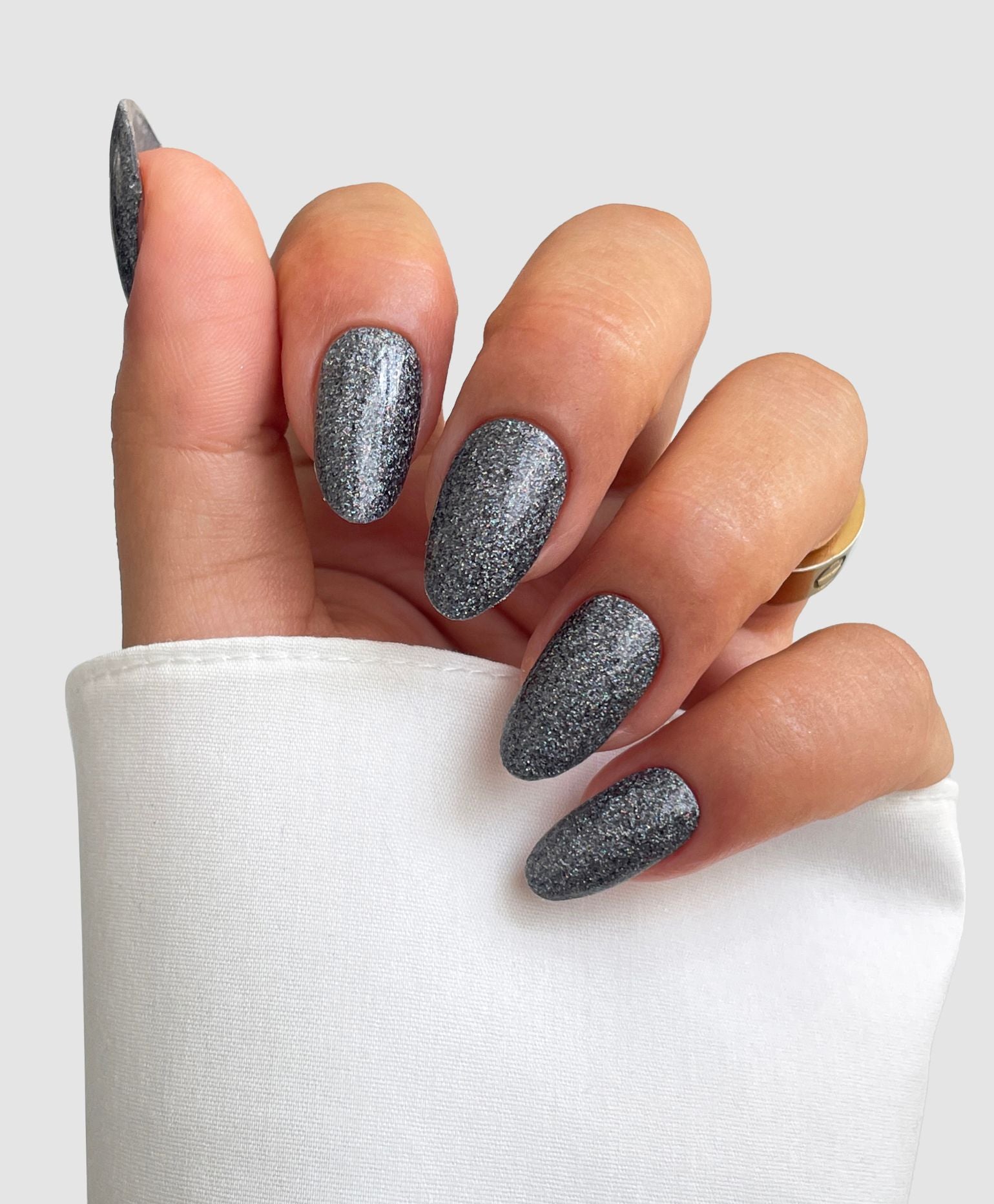 A hand with medium-length, almond-shaped nails features Sassy Saints - Brings The Salon Home’s Midnight Glimmer Gel Wrap, a sparkling silver look. The person wears a white long-sleeve top and a gold ring on their middle finger against a light background.