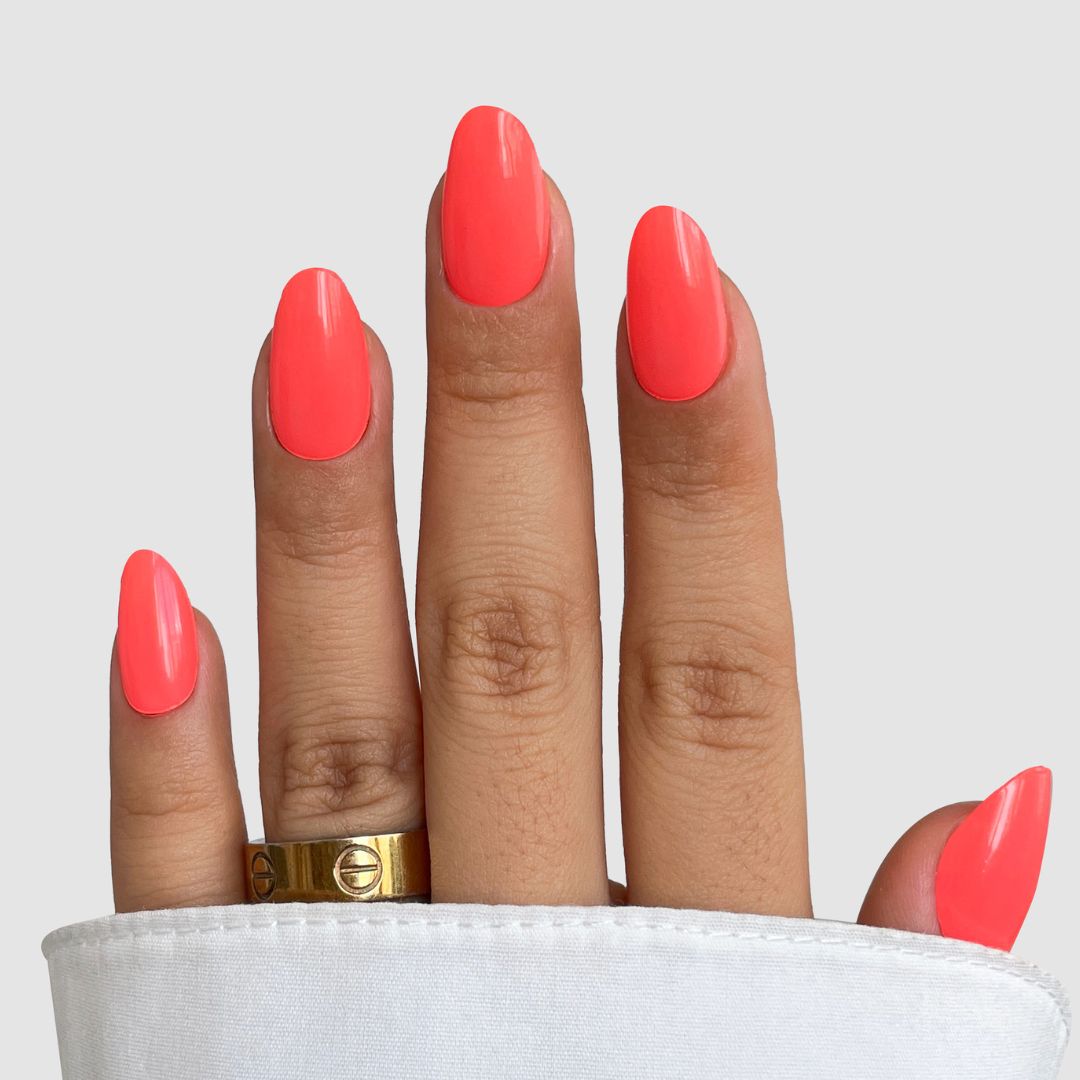 A hand with long, almond-shaped nails features Sassy Saints Coral Kiss Gel Wraps in bright coral orange, paired with a gold ring on the middle finger and a white fabric sleeve. The background is light gray.