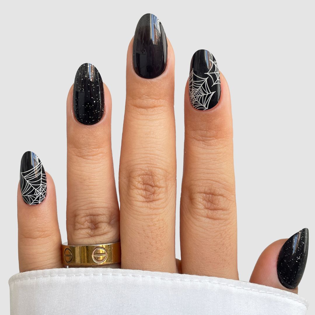A hand with black, glossy, almond-shaped Webbed Luxe Gel Wrap nails from Sassy Saints - Brings The Salon Home features white spider web designs and subtle glitter. Two gold rings adorn the ring finger, and a white sleeve partially covers the hand.