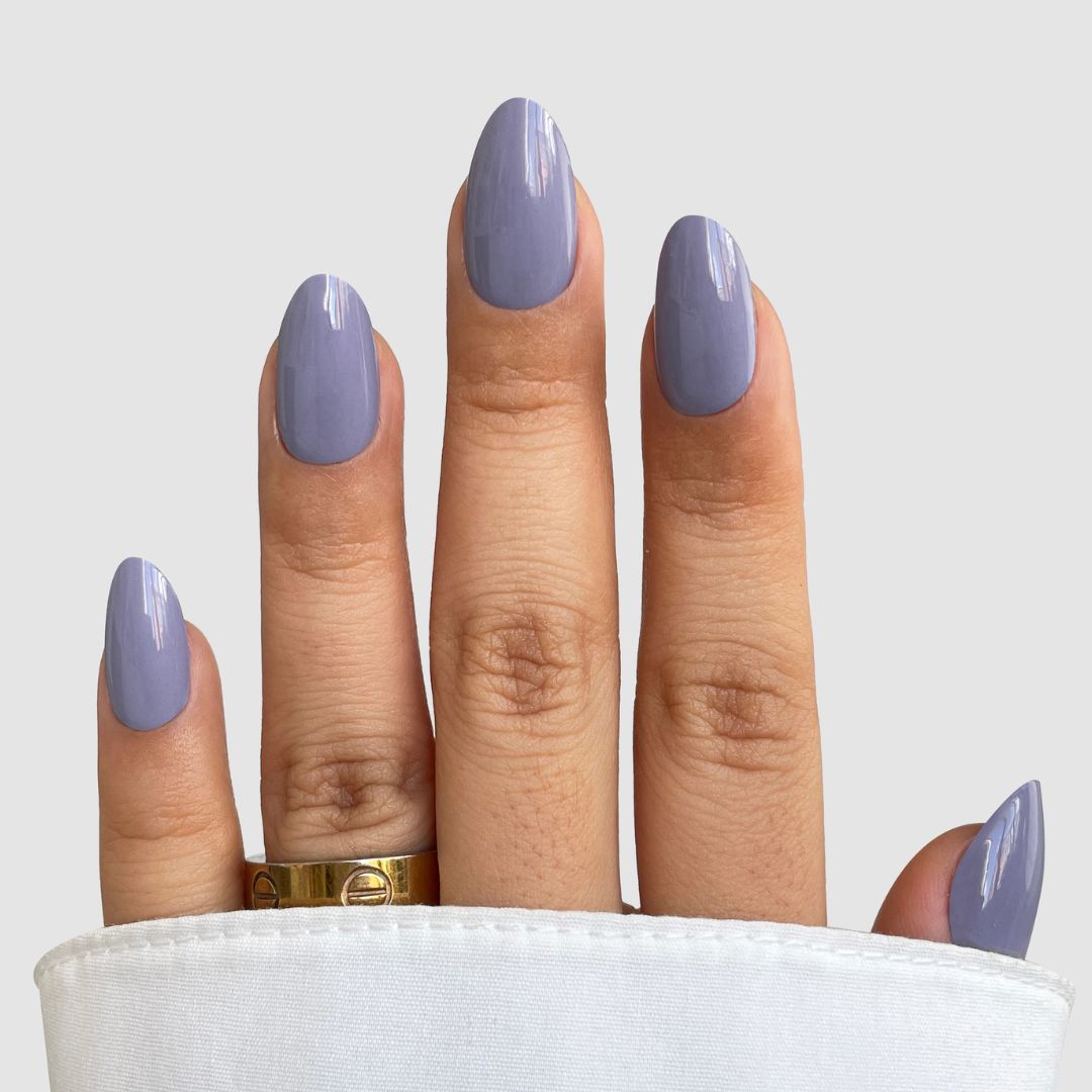 A hand with medium-length, almond-shaped nails features Sassy Saints’ Lilac Smoke Gel Wrap, a glossy lavender polish. The hand is partly covered by a white sleeve, wears a gold ring on the ring finger, and rests against a light gray background.