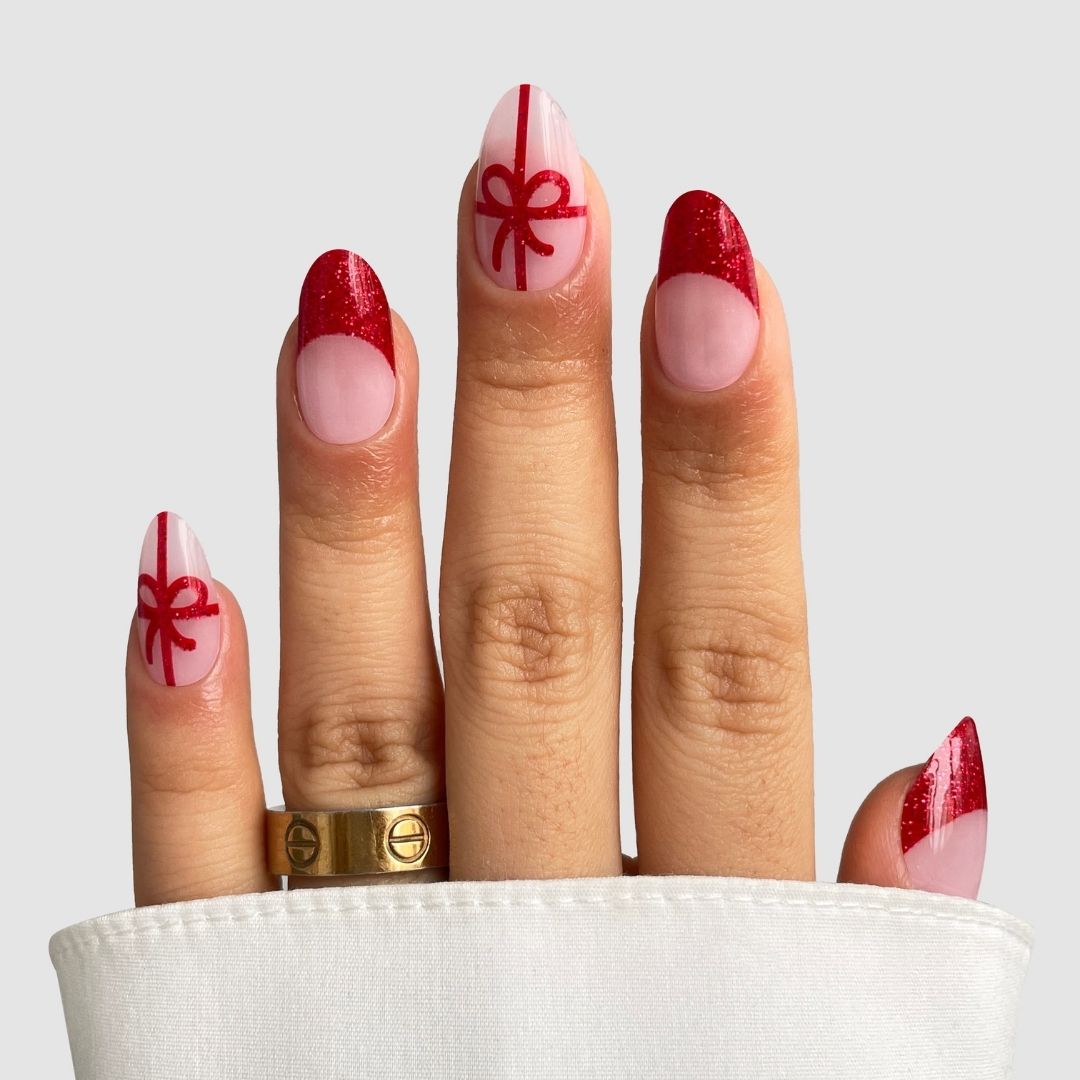 A hand wears Sassy Saints' Gift Wrapped Gel Wraps: almond nails with festive red glitter tips, ribbon and bow designs on pink and white, paired with gold rings and a white sleeve—bringing the salon home.